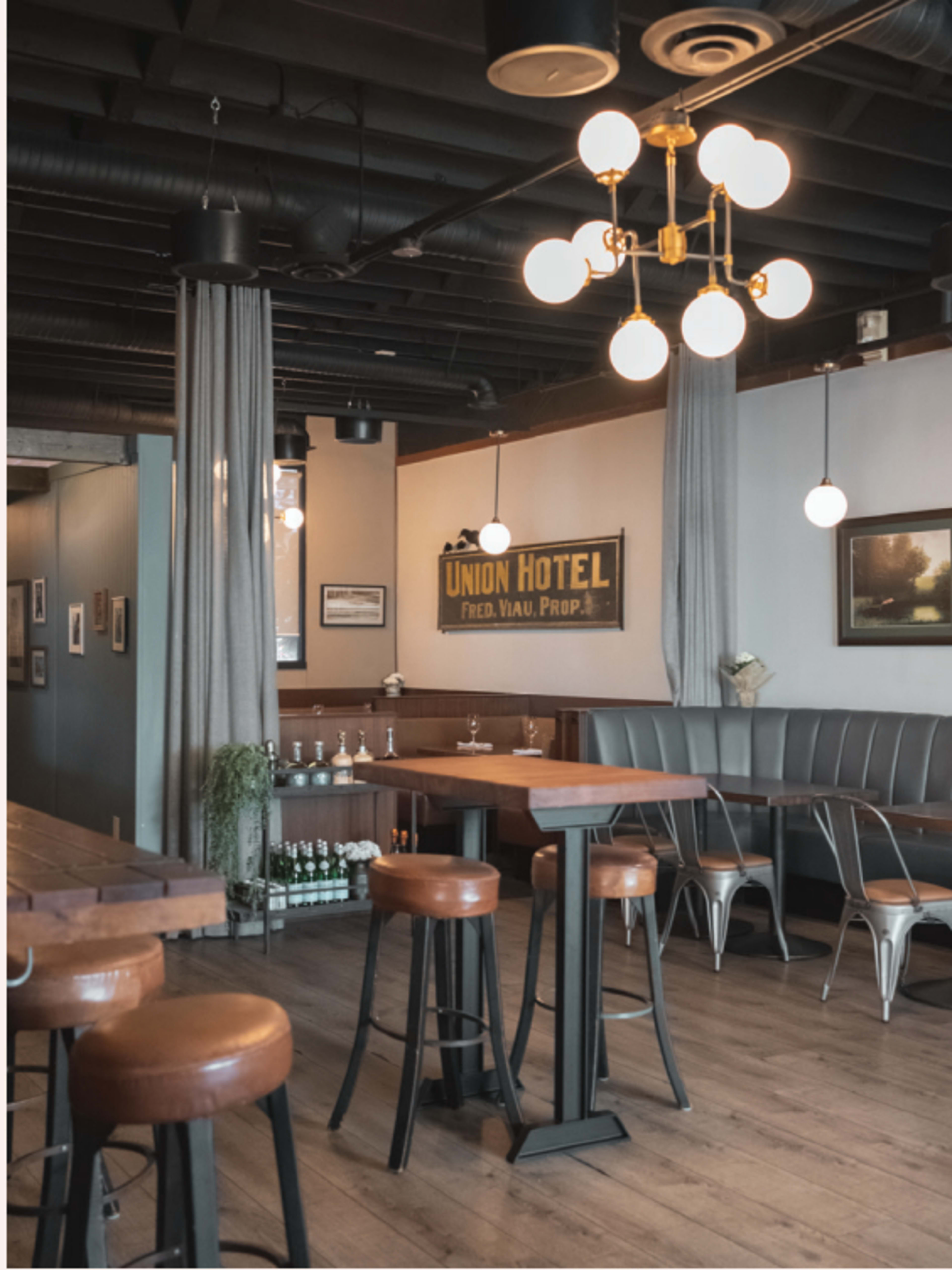 A modern bar area features wooden tables and stools, with soft lighting and gray curtains against a backdrop of framed pictures and a vintage sign.