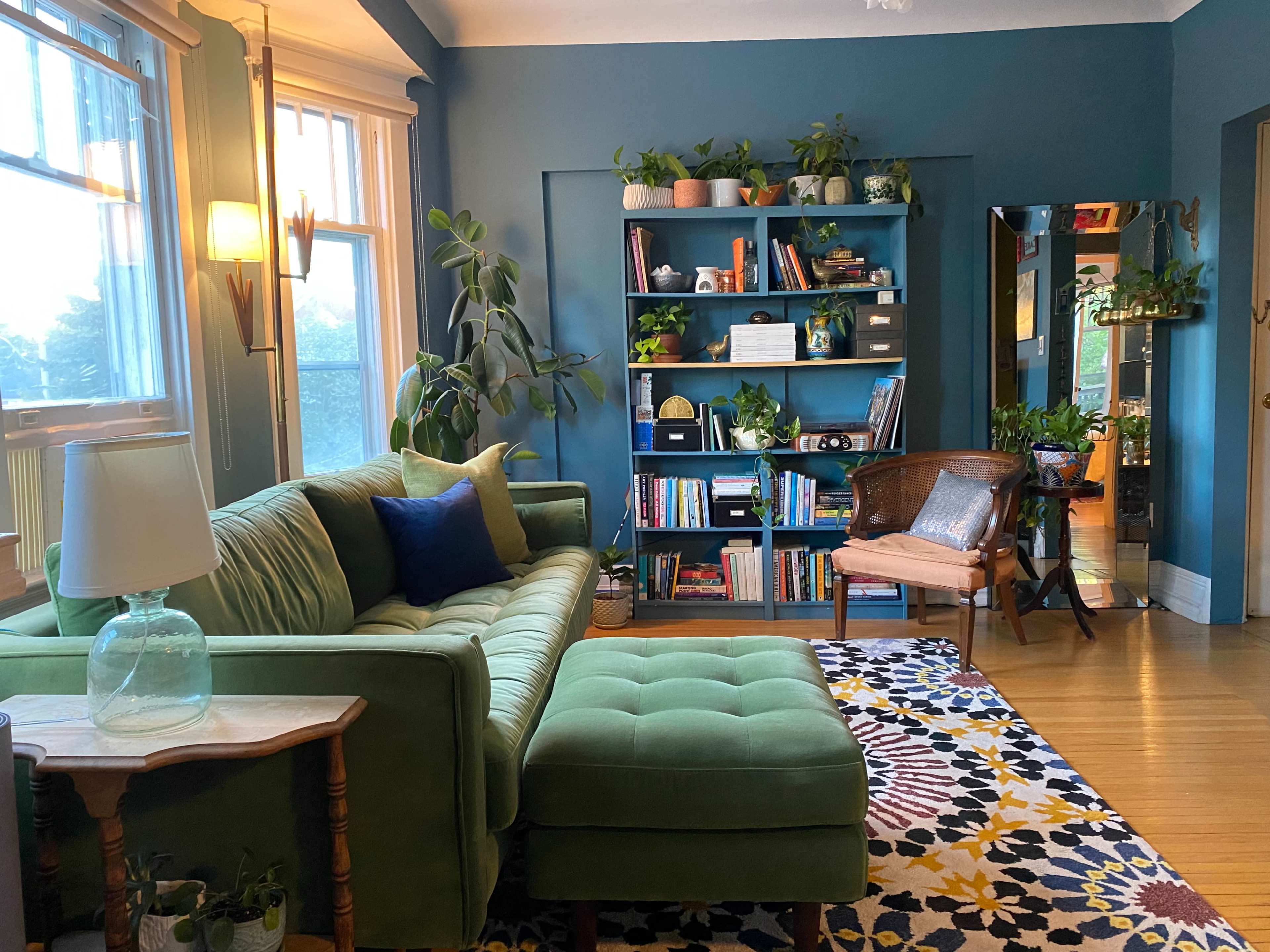 A cozy living room featuring a green sofa, a bookshelf filled with plants and books, and a patterned rug on a hardwood floor.