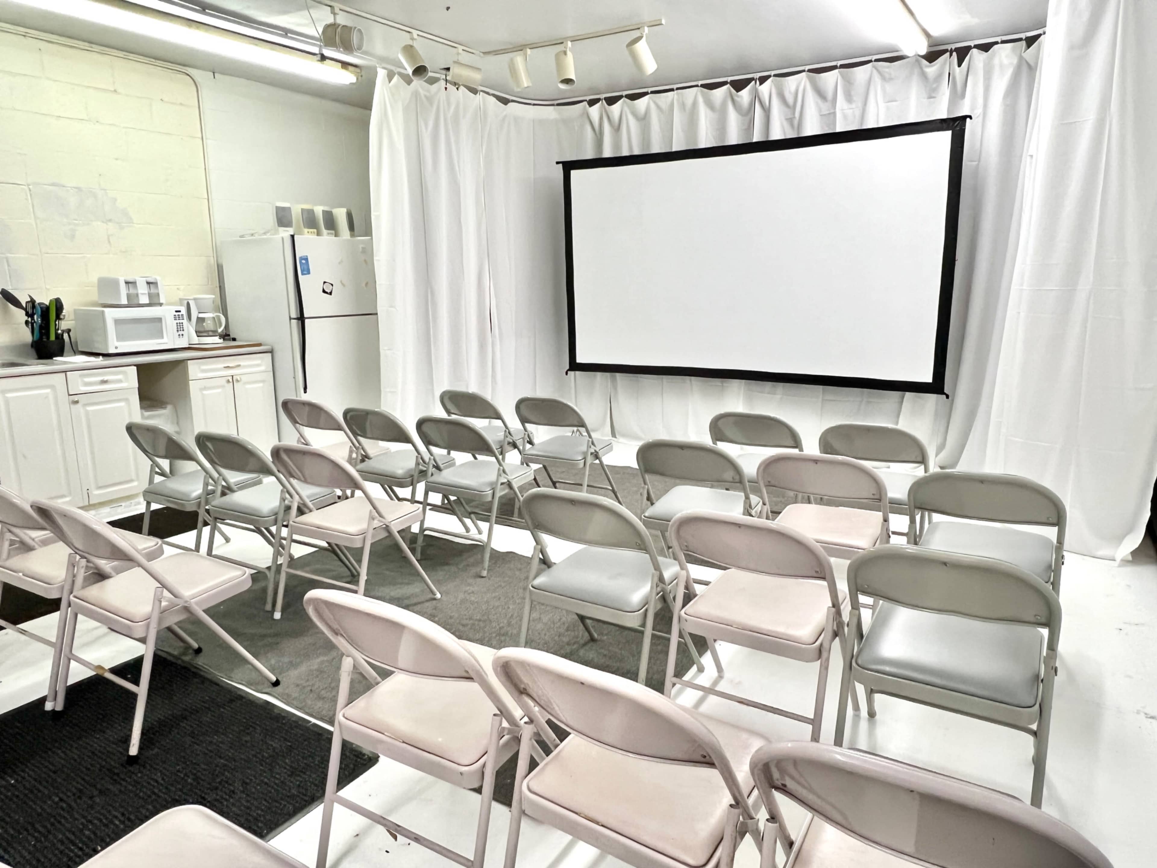 The image shows a small room with a projection screen at the front, arranged with several rows of folding chairs and a kitchenette area in the corner.
