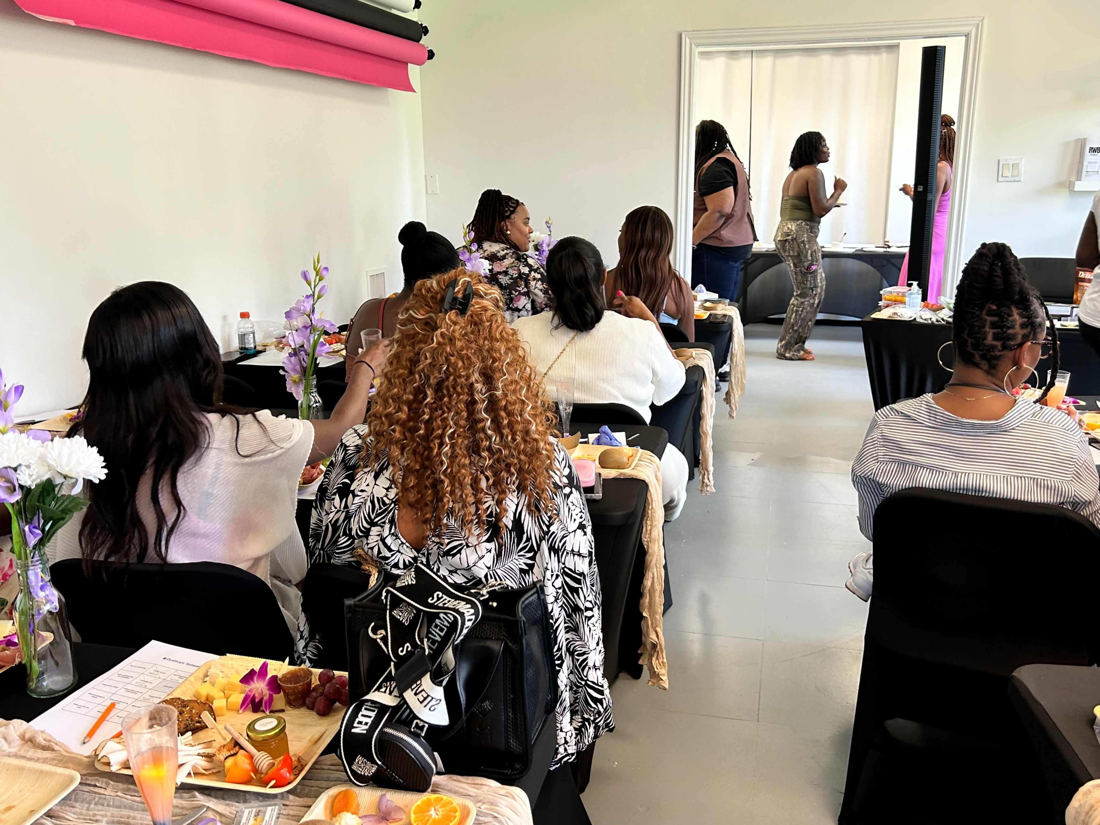A group of people sits at tables in a brightly lit room, with food and drinks placed in front of them, while others socialize in the background.