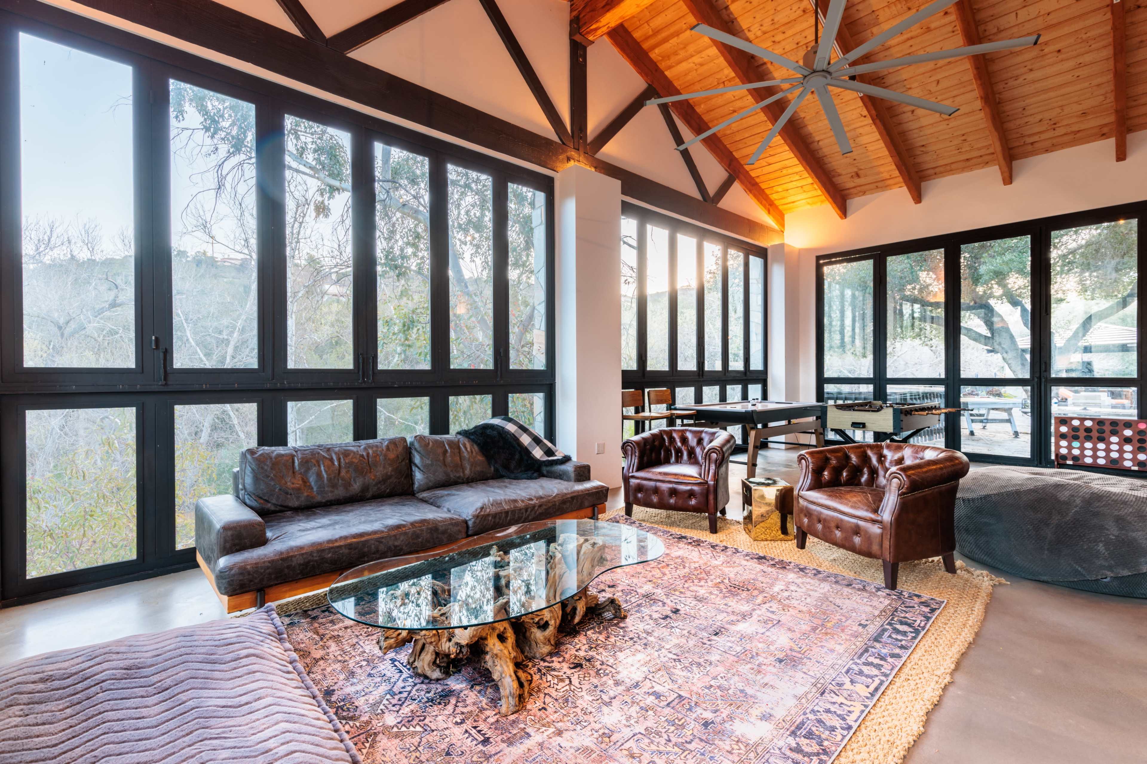 The image shows a modern living room with large windows, a leather sofa, two armchairs, a wooden coffee table, and a patterned rug.