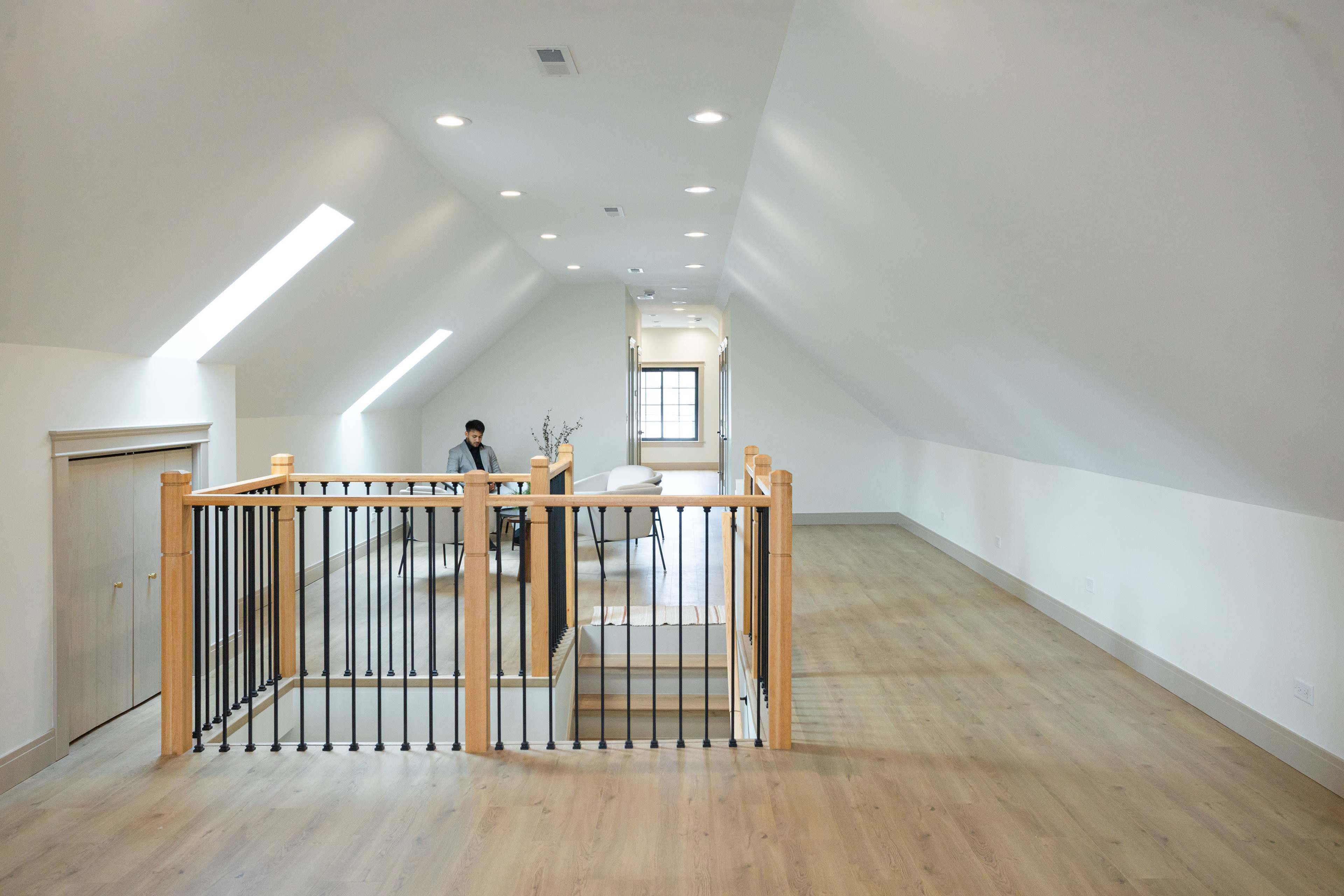 The image shows a spacious attic space with wooden flooring, sloped ceilings, and a railing overlooking a lower area furnished with a table and chair.