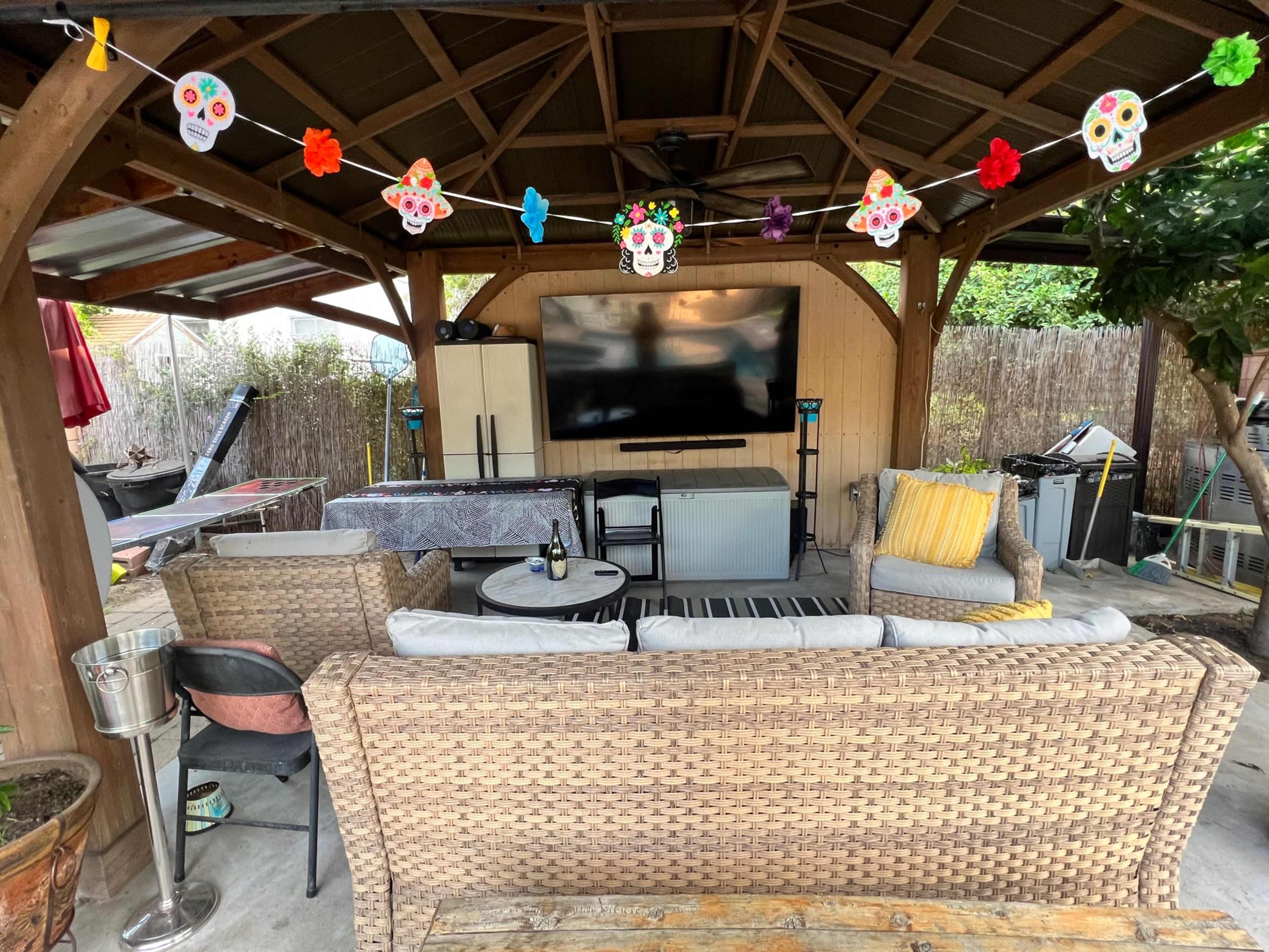 A covered outdoor seating area features a television mounted on the wall, a table, and various chairs, with colorful decorations hanging overhead.