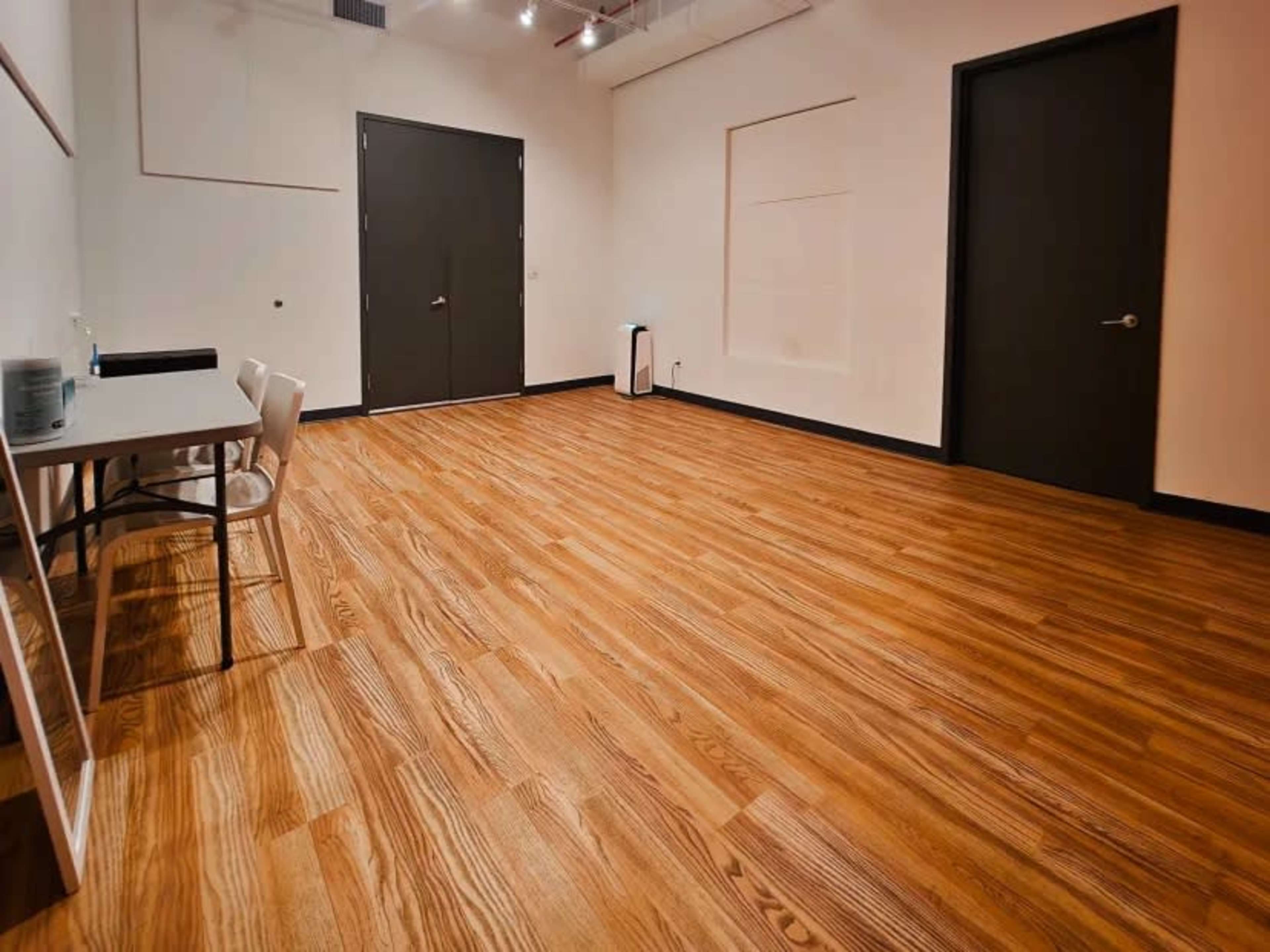 The image shows a spacious, empty room with wooden floors, two doors, a white table, and a chair against one wall.