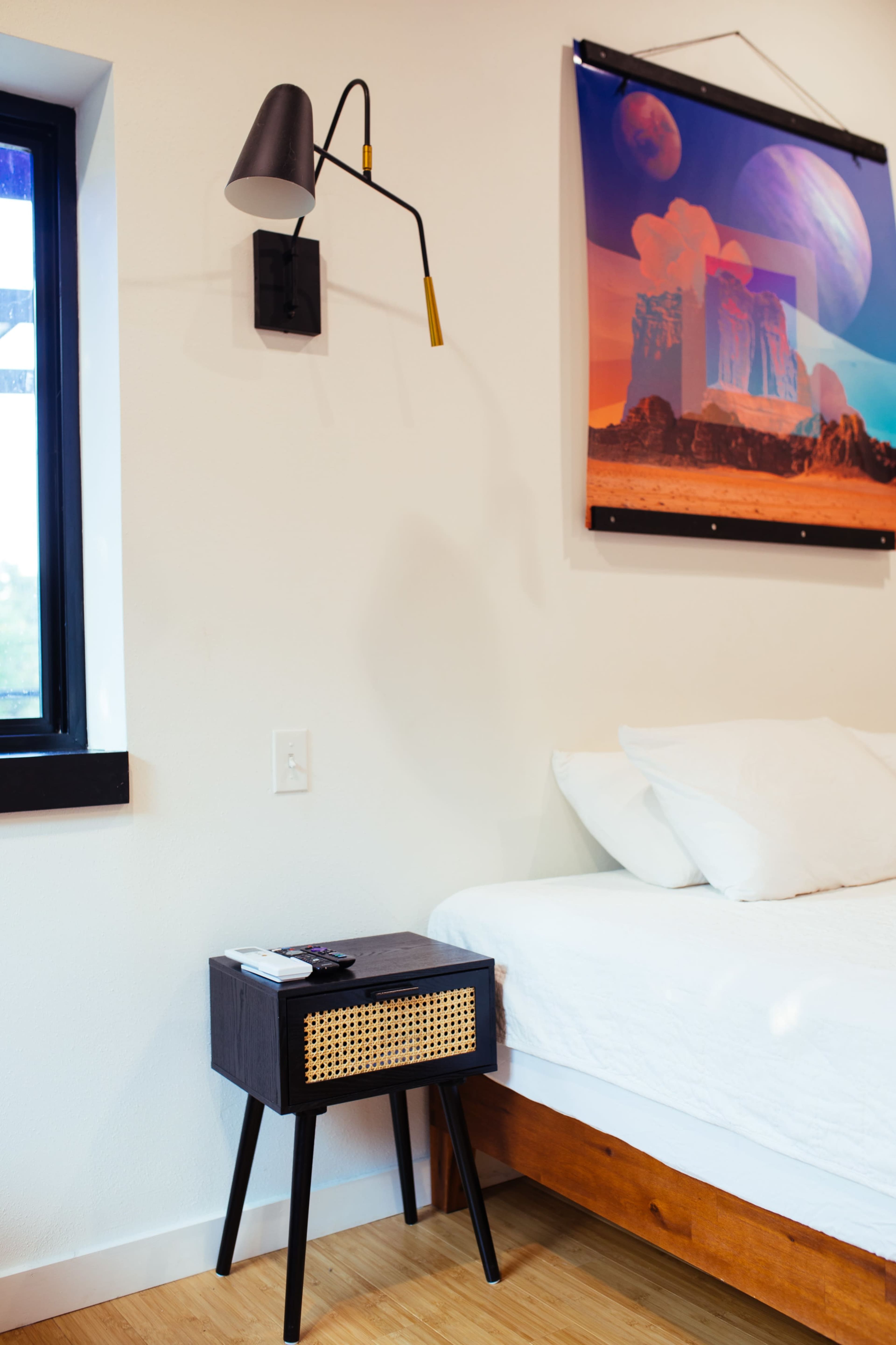 A minimalist bedroom featuring a bed with white bedding, a small nightstand with a remote, and a colorful abstract artwork on the wall above it.