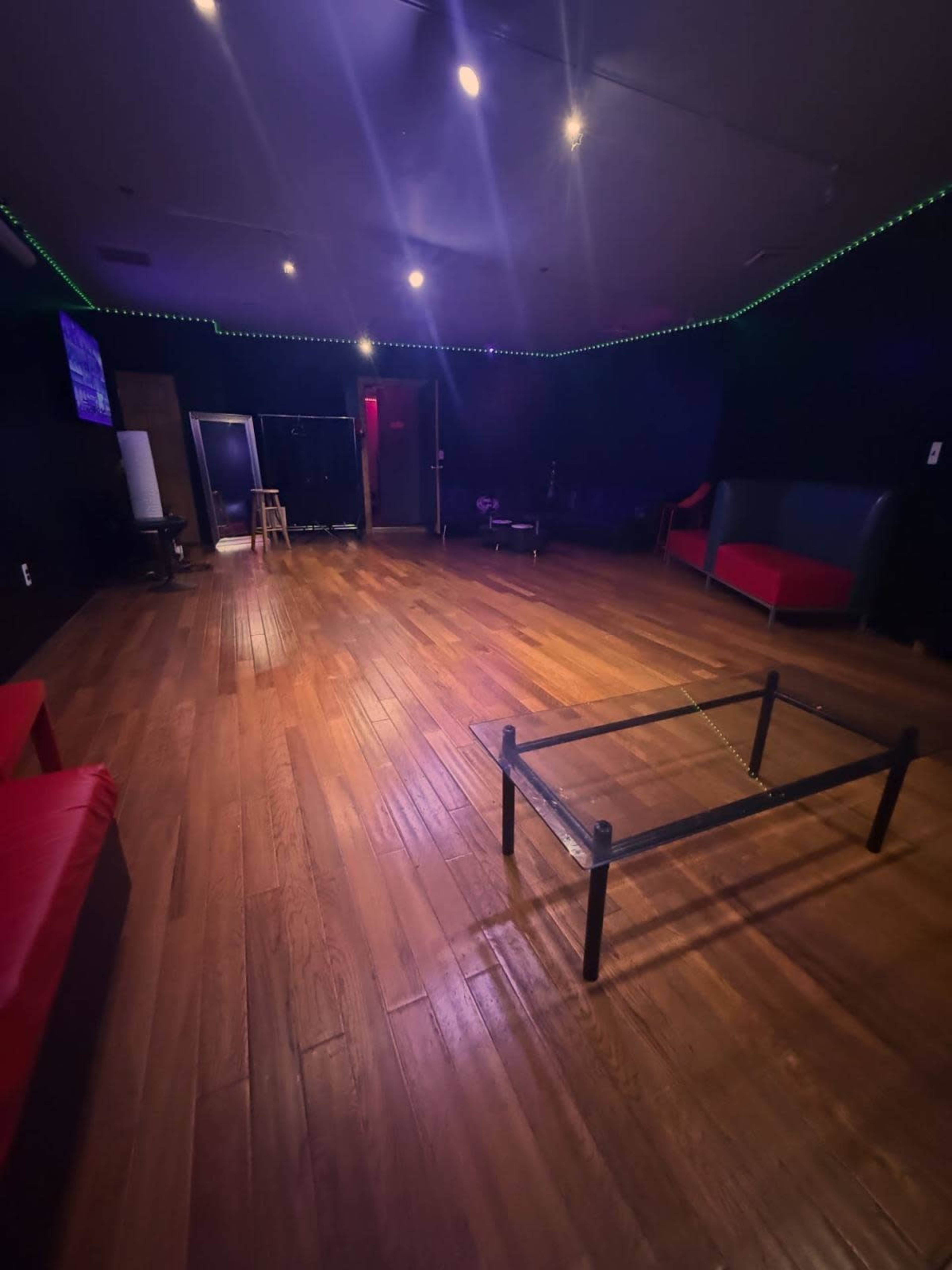The image shows a dimly lit interior room with dark walls, hardwood flooring, a glass coffee table in the center, red seating along the perimeter, and a small stool in one corner.