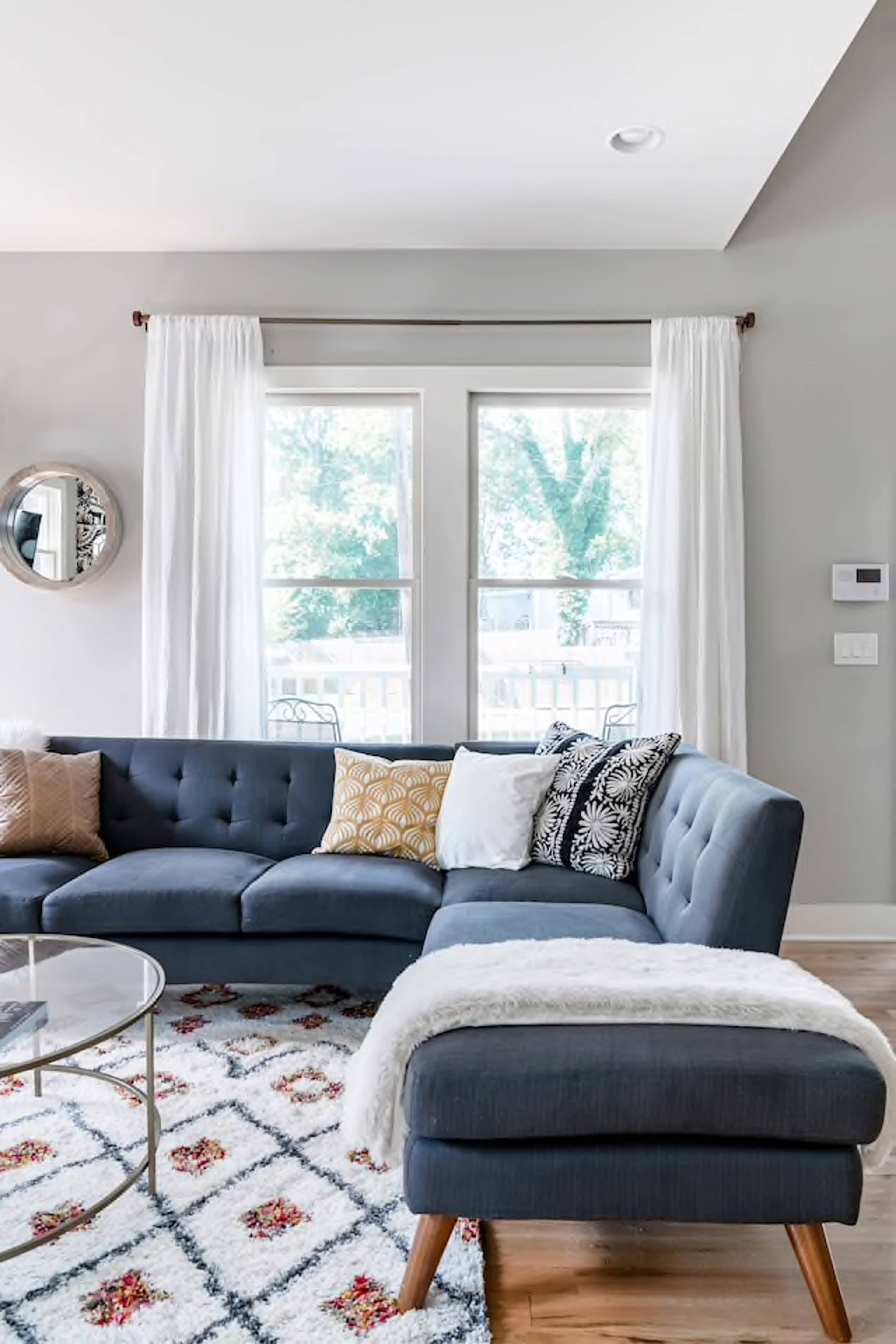 A modern living room with a sectional sofa, a round glass coffee table, and large windows dressed with white curtains.