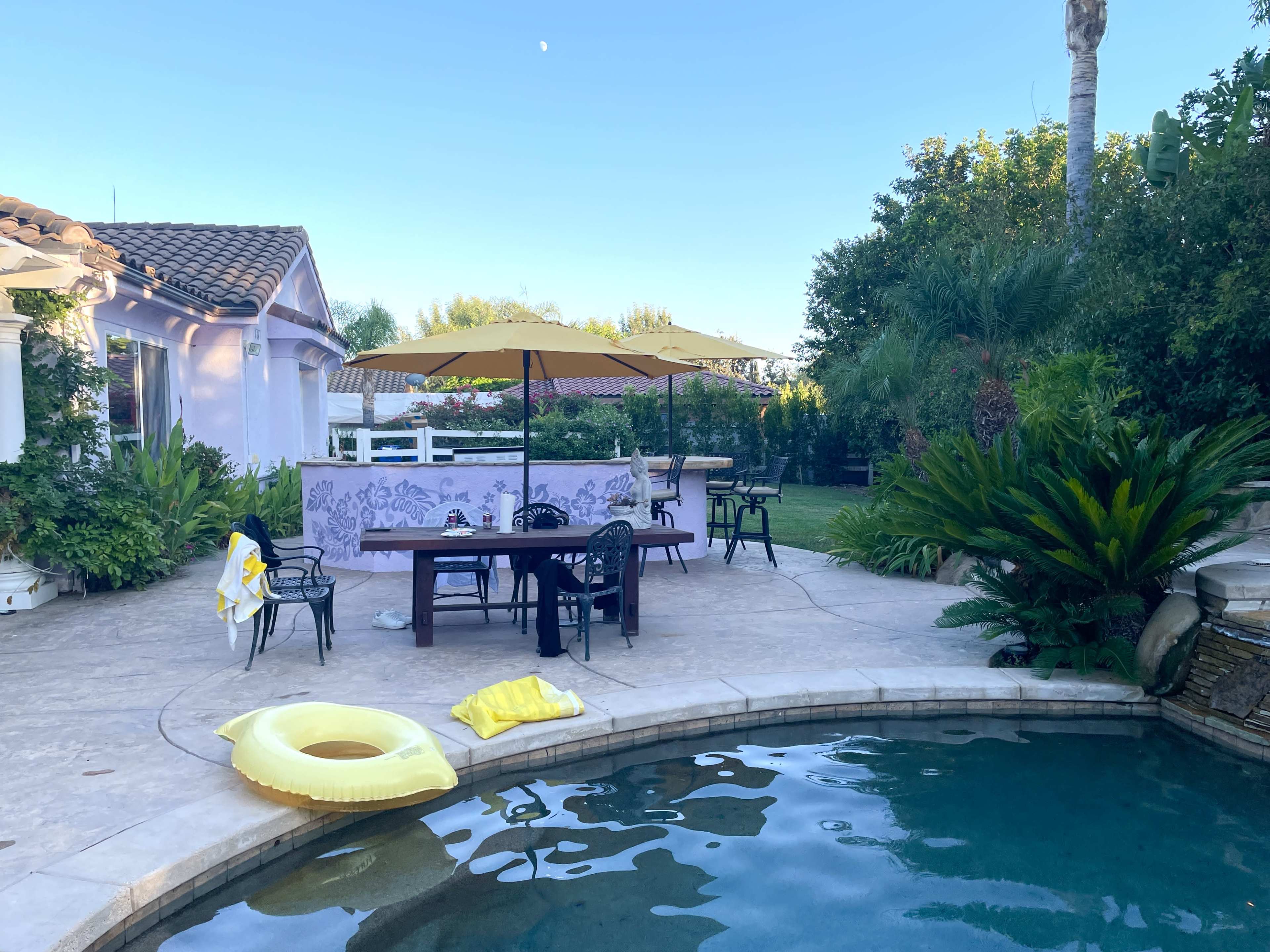 A backyard pool area features a round pool, a dining table with yellow umbrellas, and a few lounge chairs, alongside a grassy lawn and greenery.