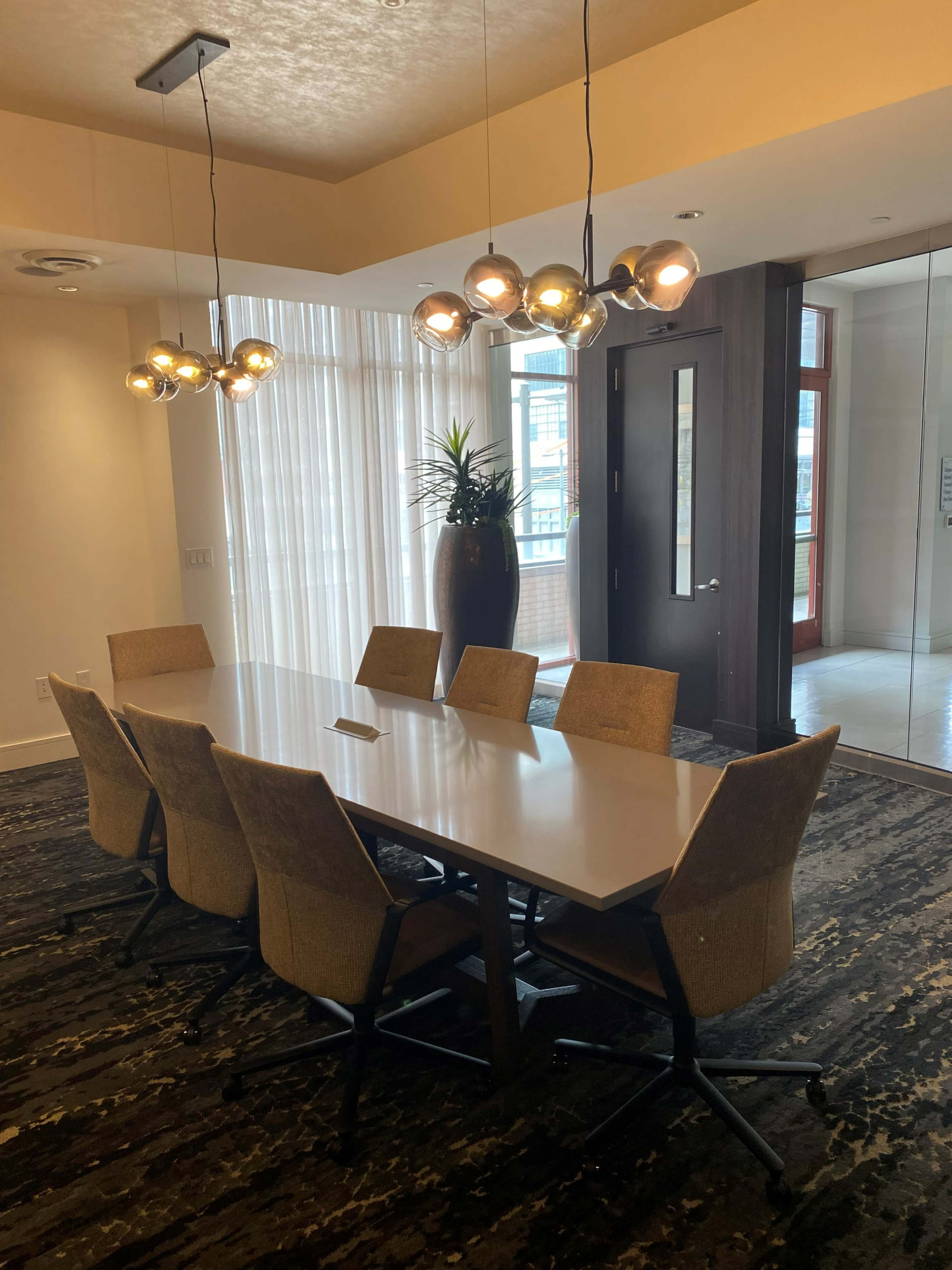A conference room features a large table surrounded by chairs, with a decorative plant in the corner and modern lighting overhead.
