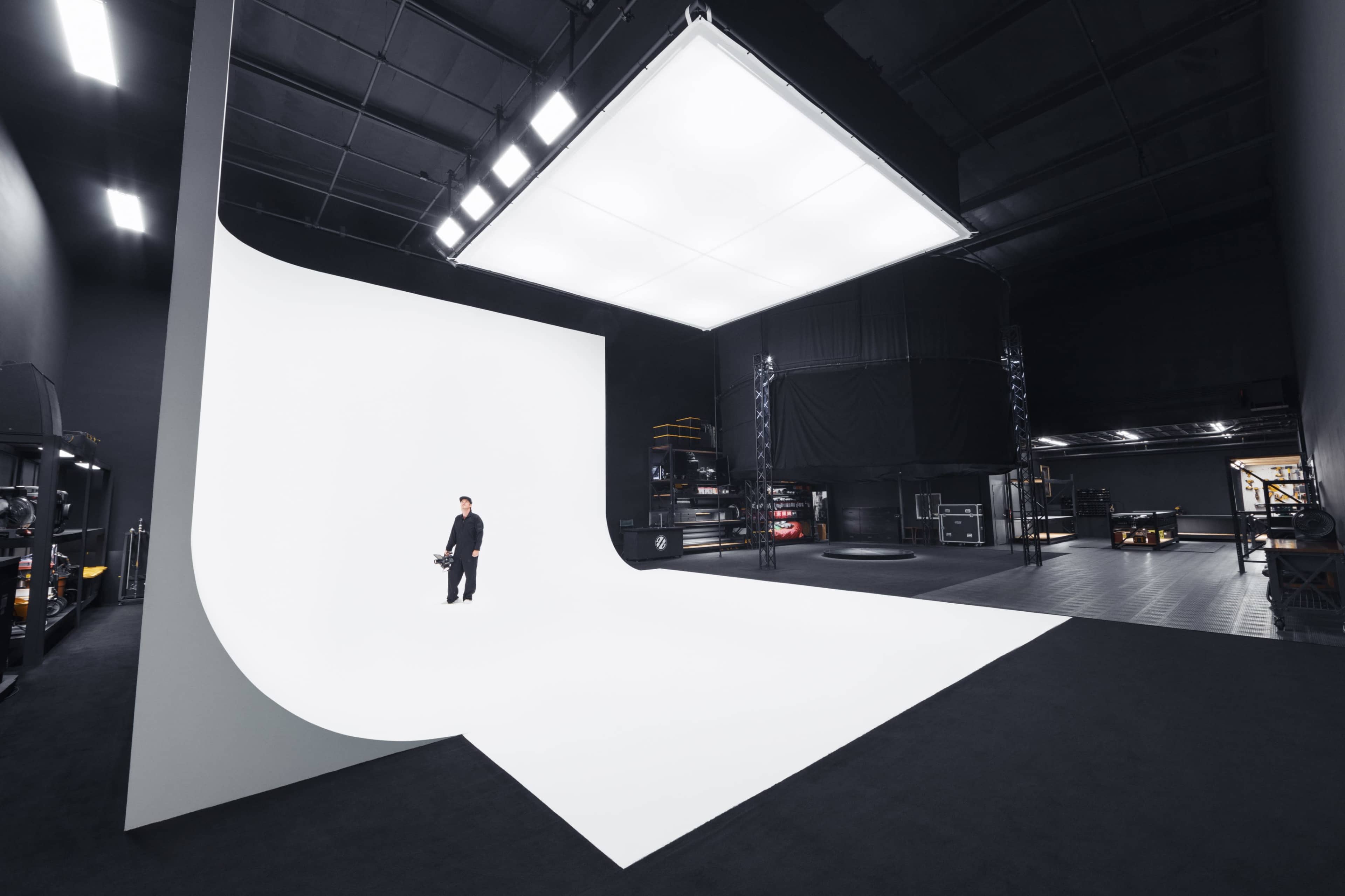 A person stands on a large white backdrop inside a modern studio space with black walls and various equipment visible in the background.