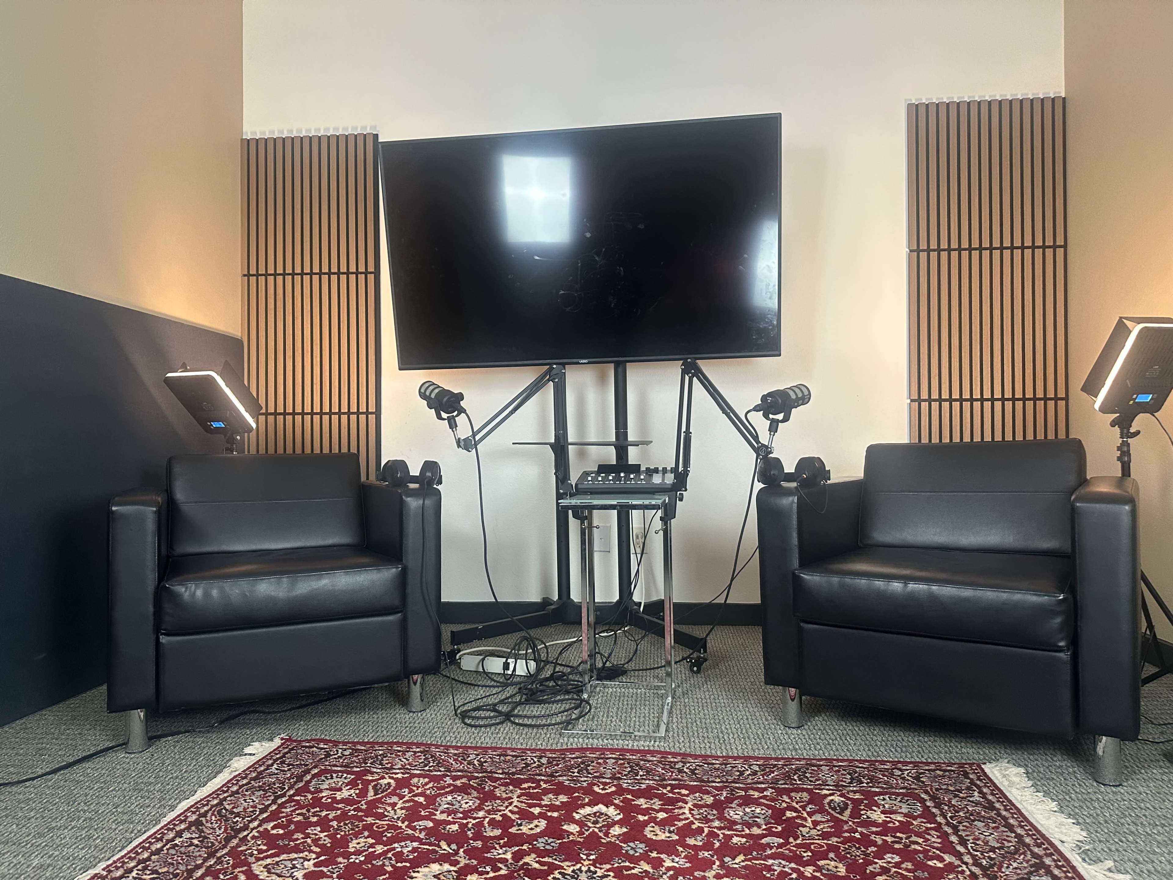 The image shows a recording studio setup featuring two black leather chairs facing a large screen, with microphones and lighting equipment positioned on stands in front of a patterned rug.