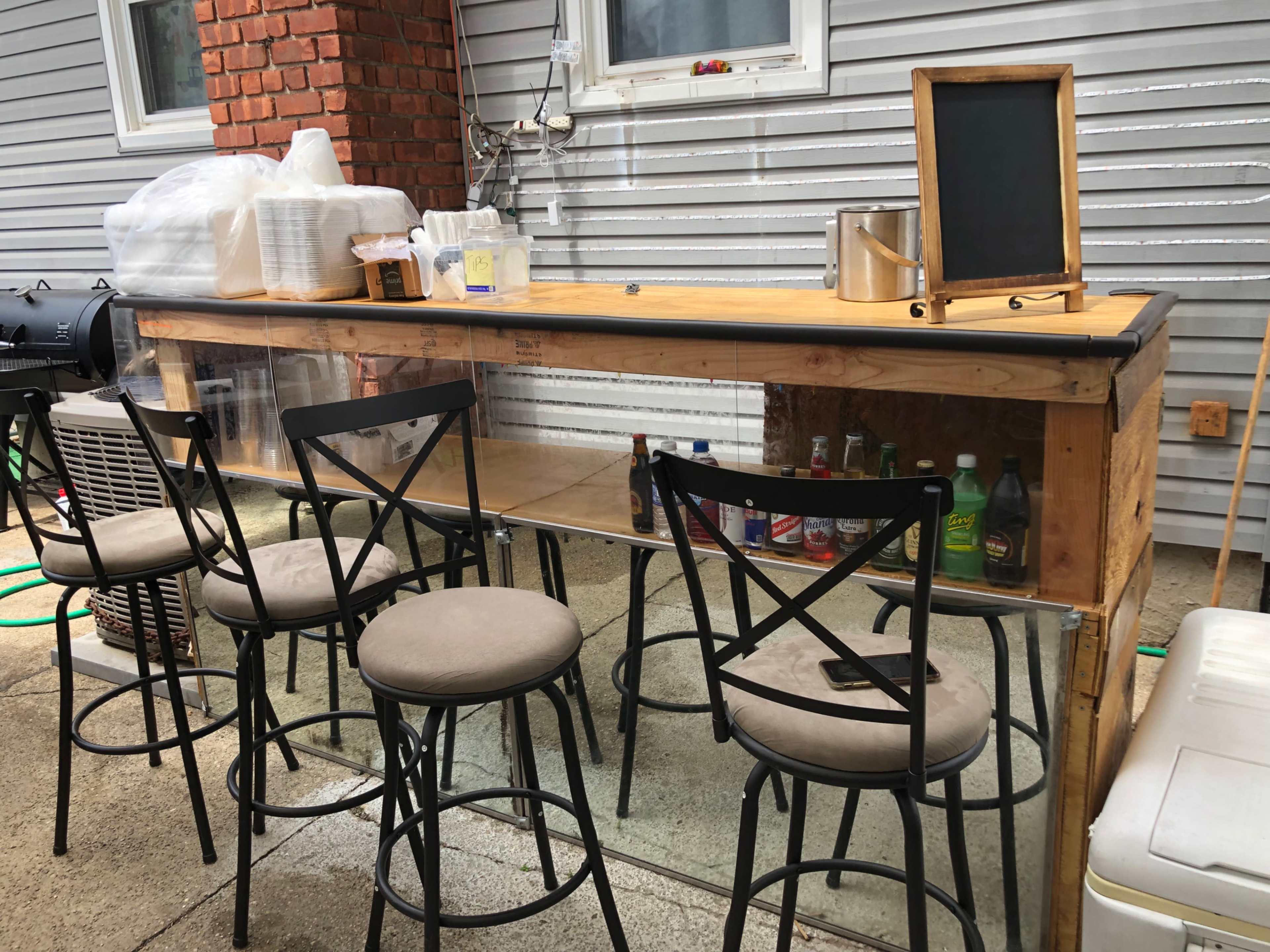 An outdoor bar area features a wooden countertop with transparent sides and several black metal and cushioned stools positioned around it.