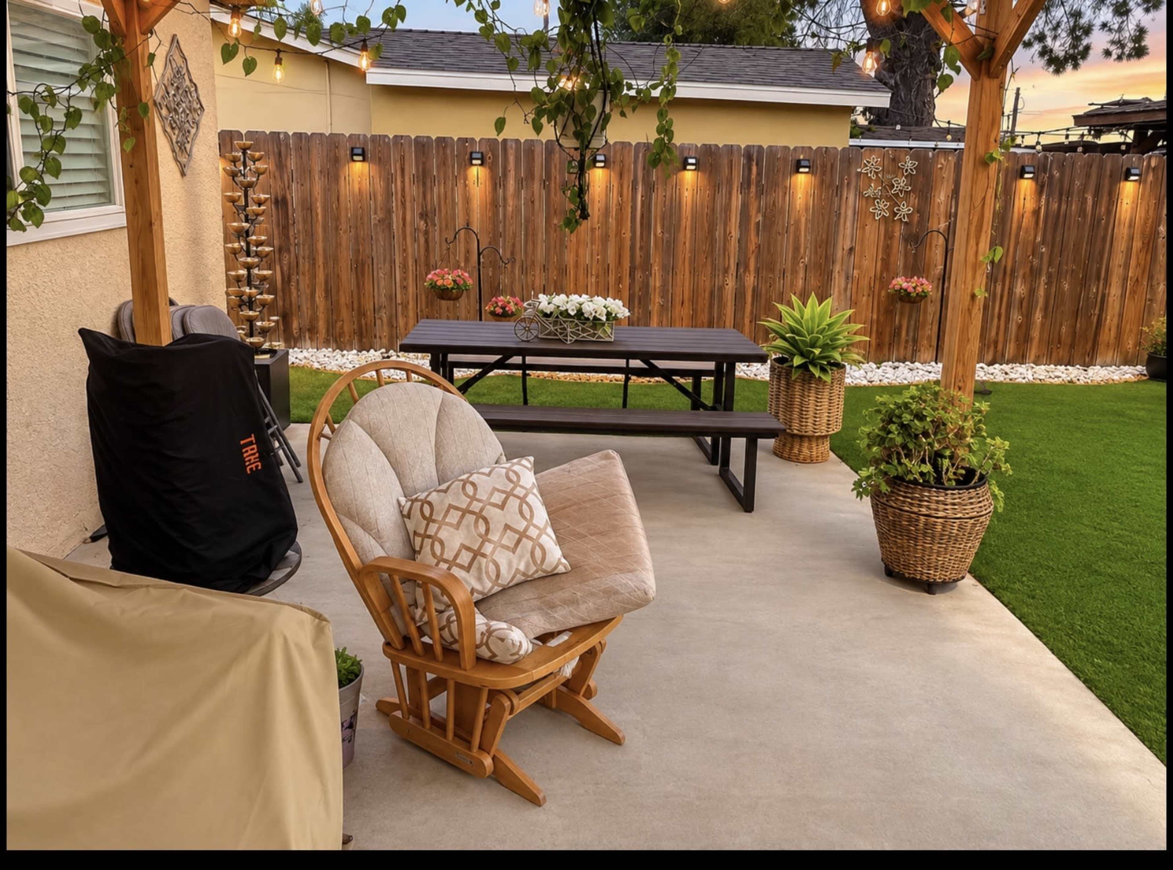 The image shows a patio area with a rocking chair, a picnic table, potted plants, and decorative lighting along a wooden fence.