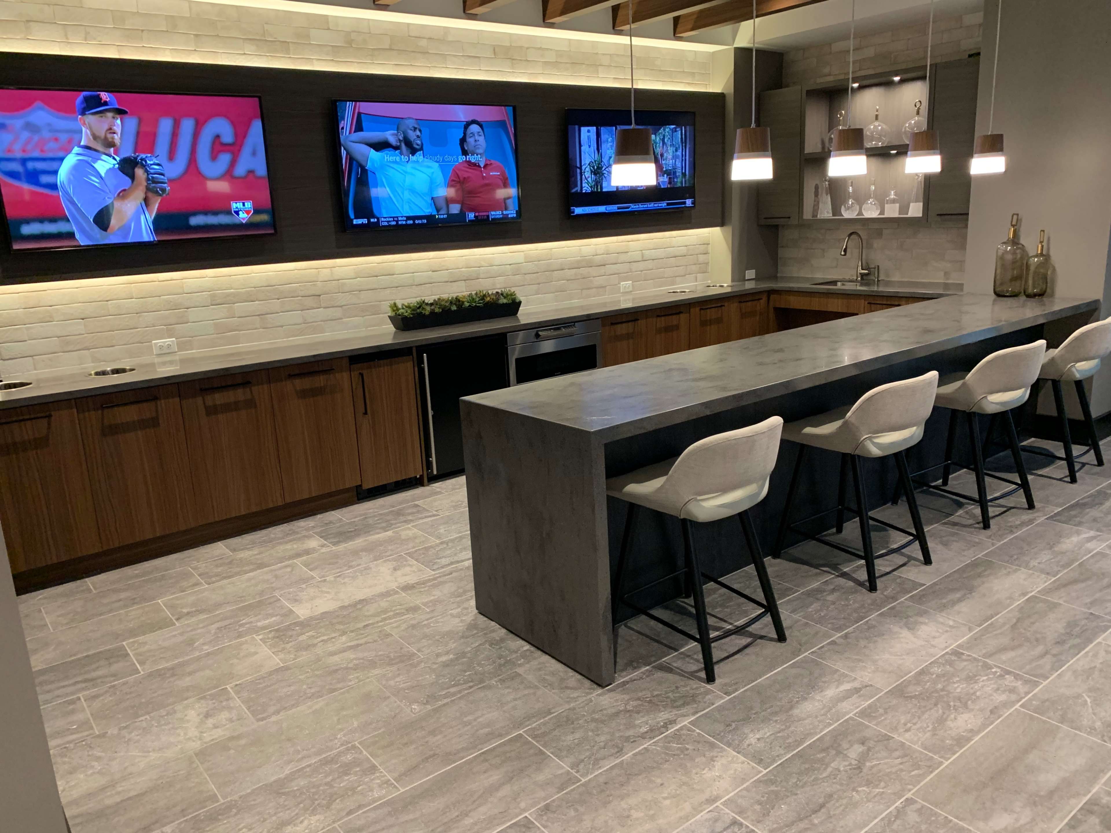 A modern kitchen bar area with a long concrete counter, eight high chairs, and three televisions mounted on the wall displaying sports content.