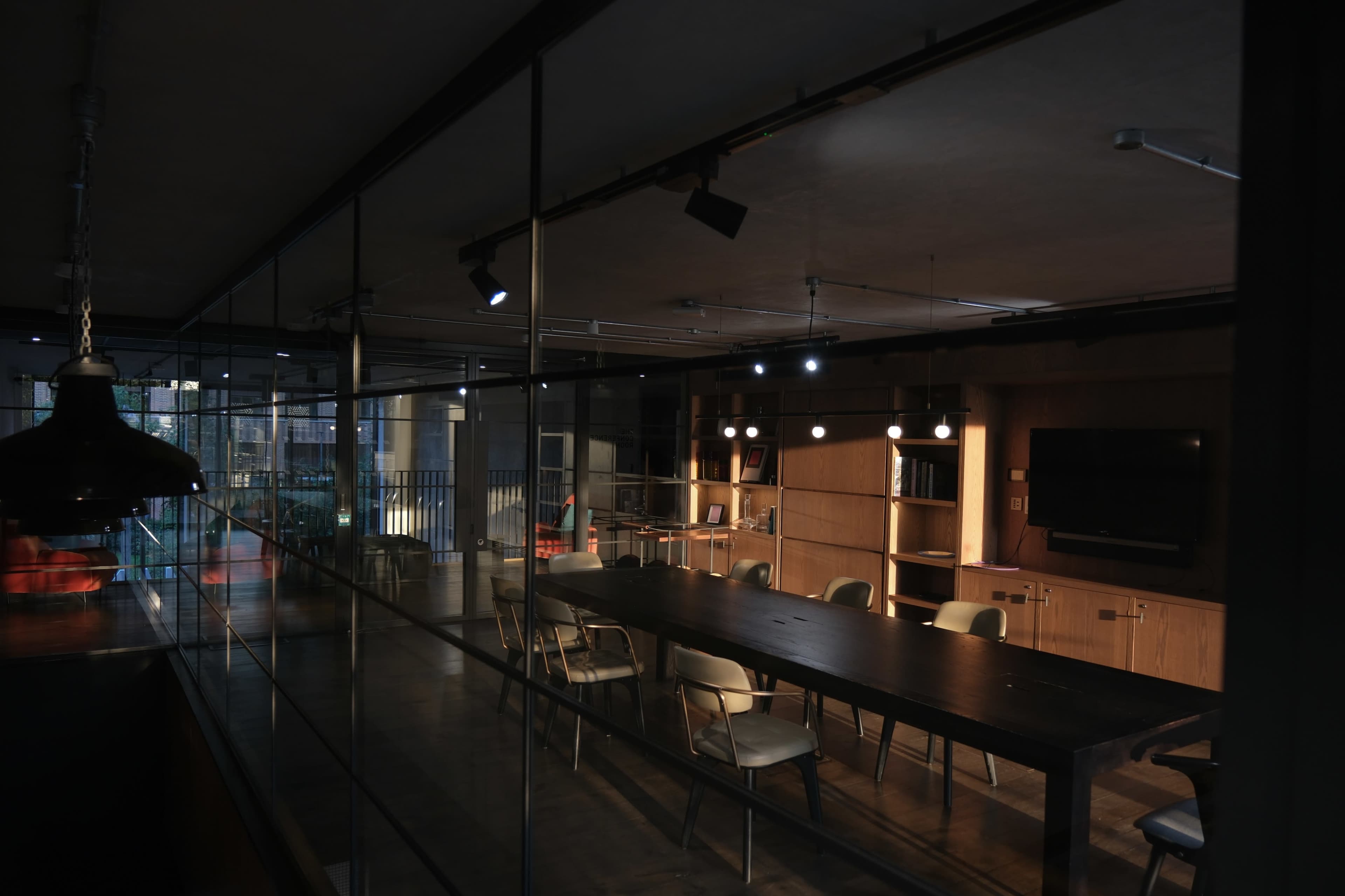 A modern meeting room with a long wooden table surrounded by white chairs, softly lit by overhead lights in a minimalist setting.