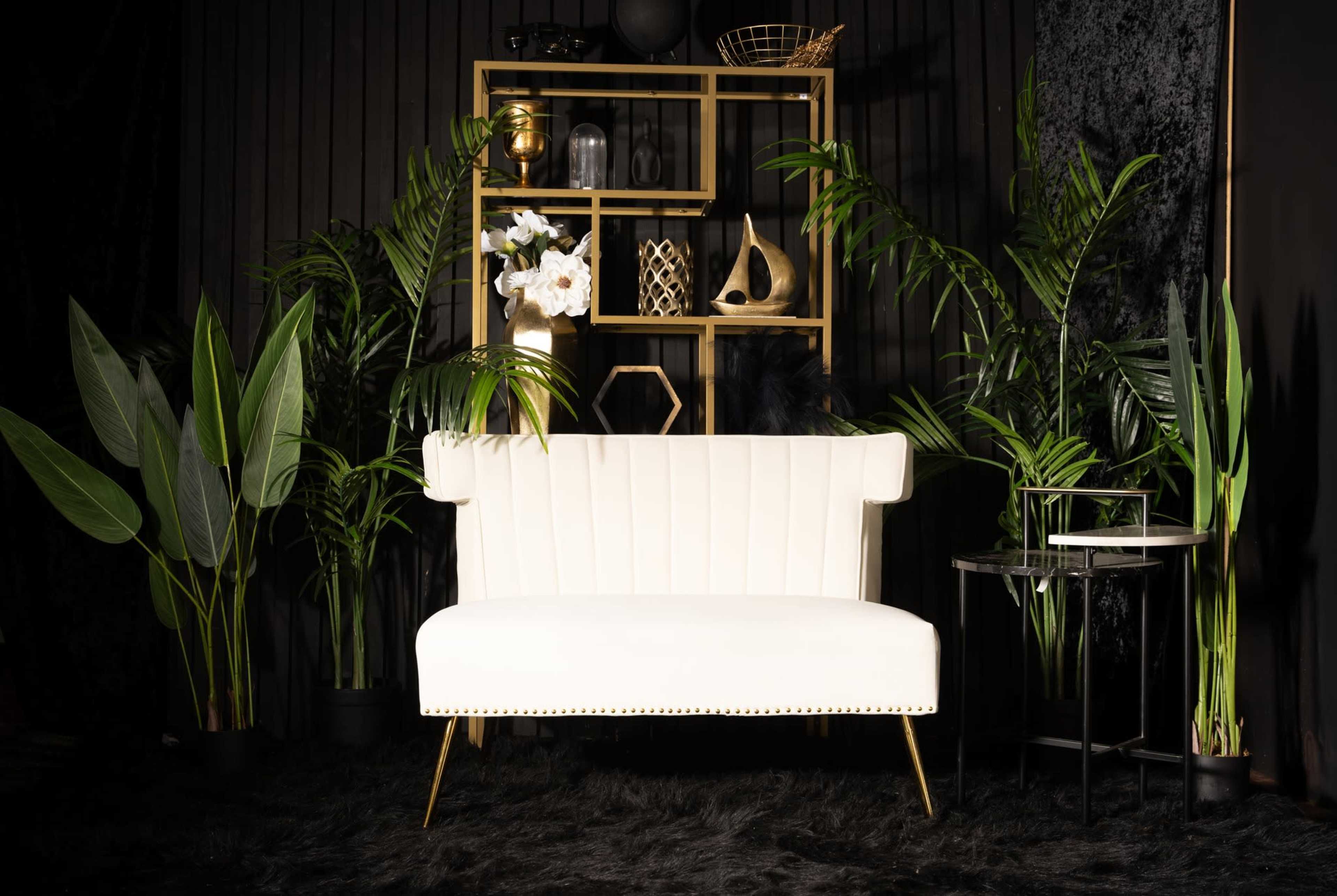 The image features a white tufted sofa in front of a gold shelving unit filled with decorative items, surrounded by large green plants against a dark background.