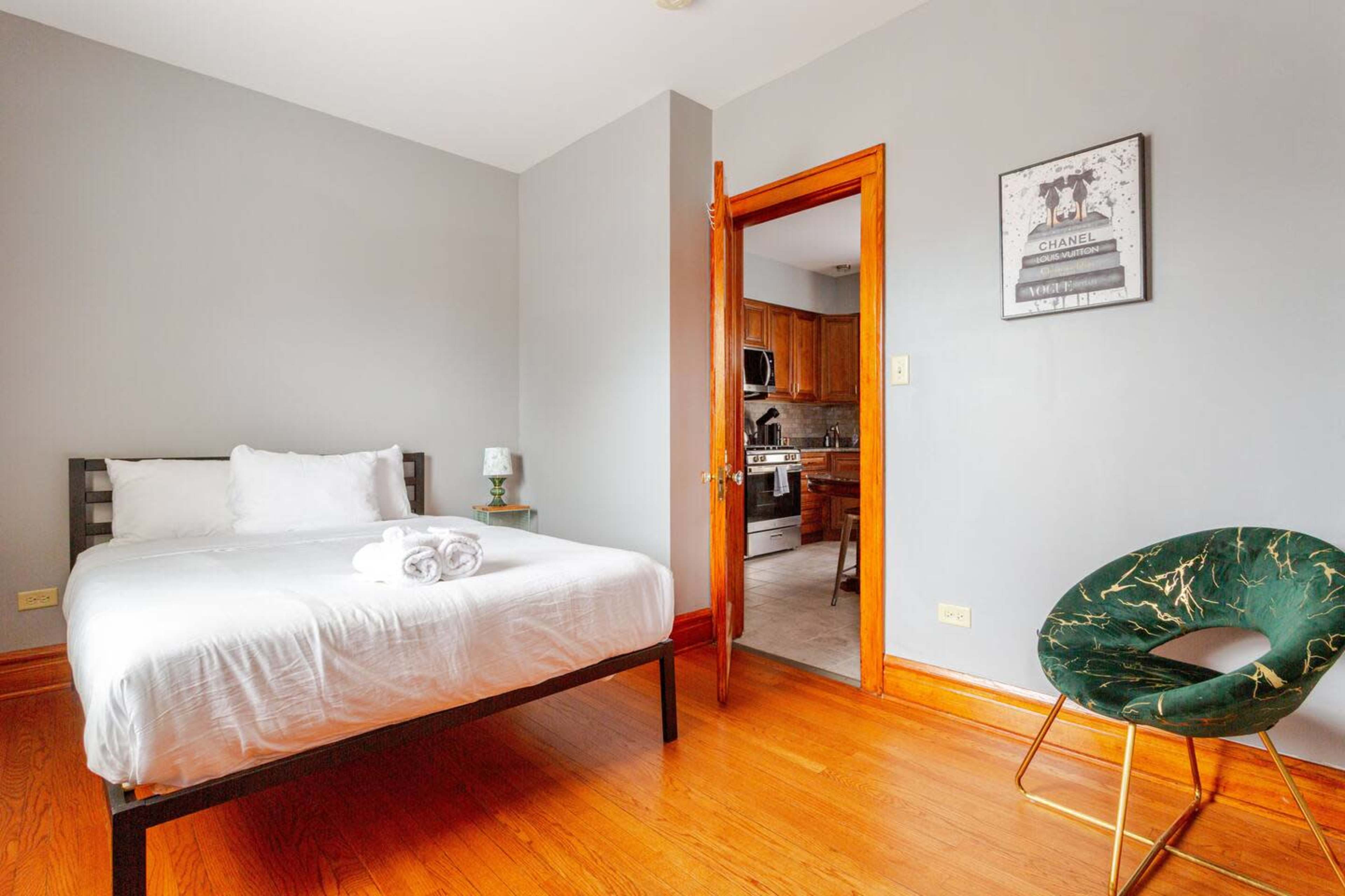 The image shows a simple bedroom with a bed, a green chair, and an open door leading to a kitchen area.