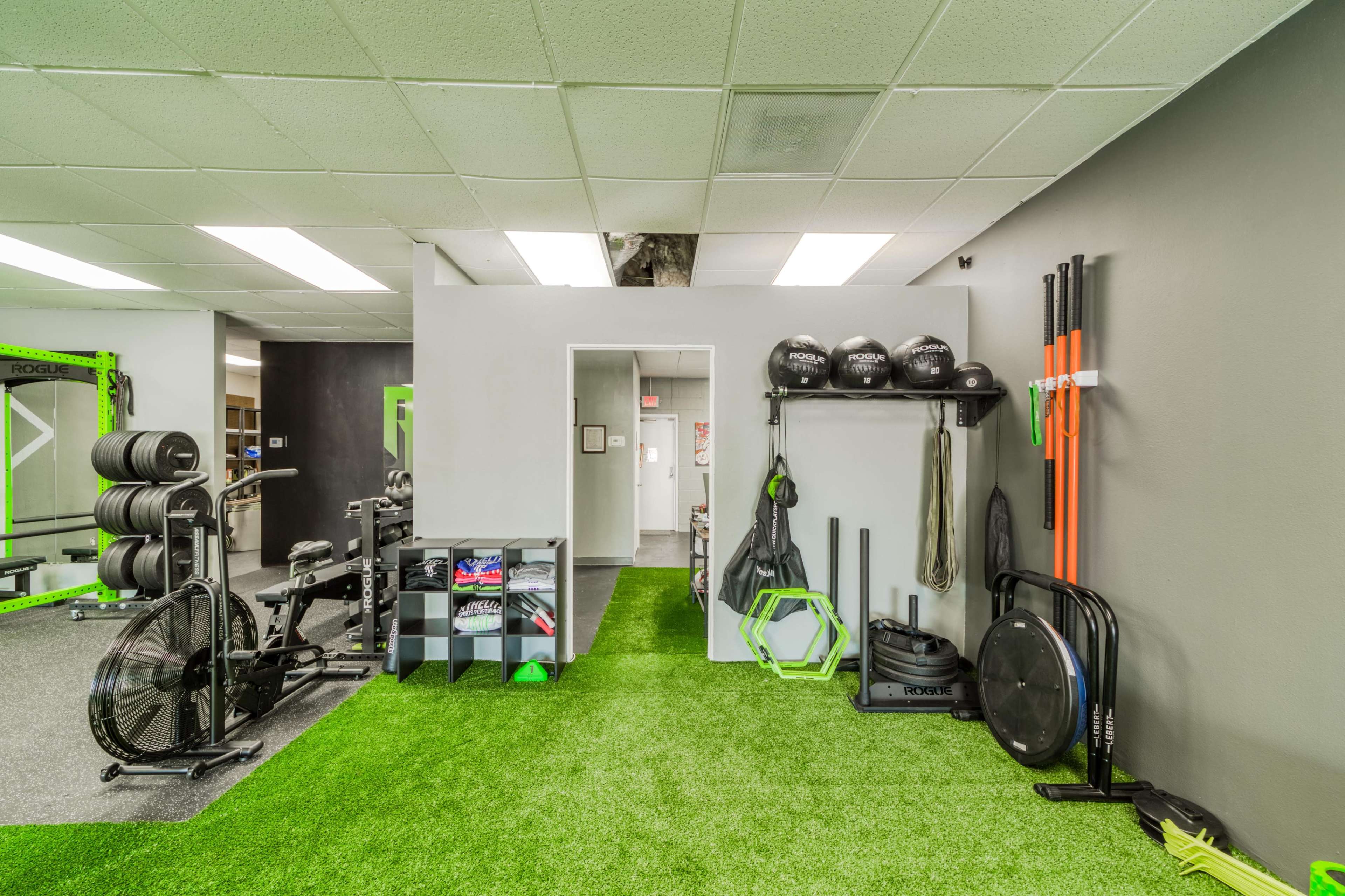 A gym interior with exercise equipment, including free weights, a rowing machine, and various fitness accessories on artificial grass flooring.