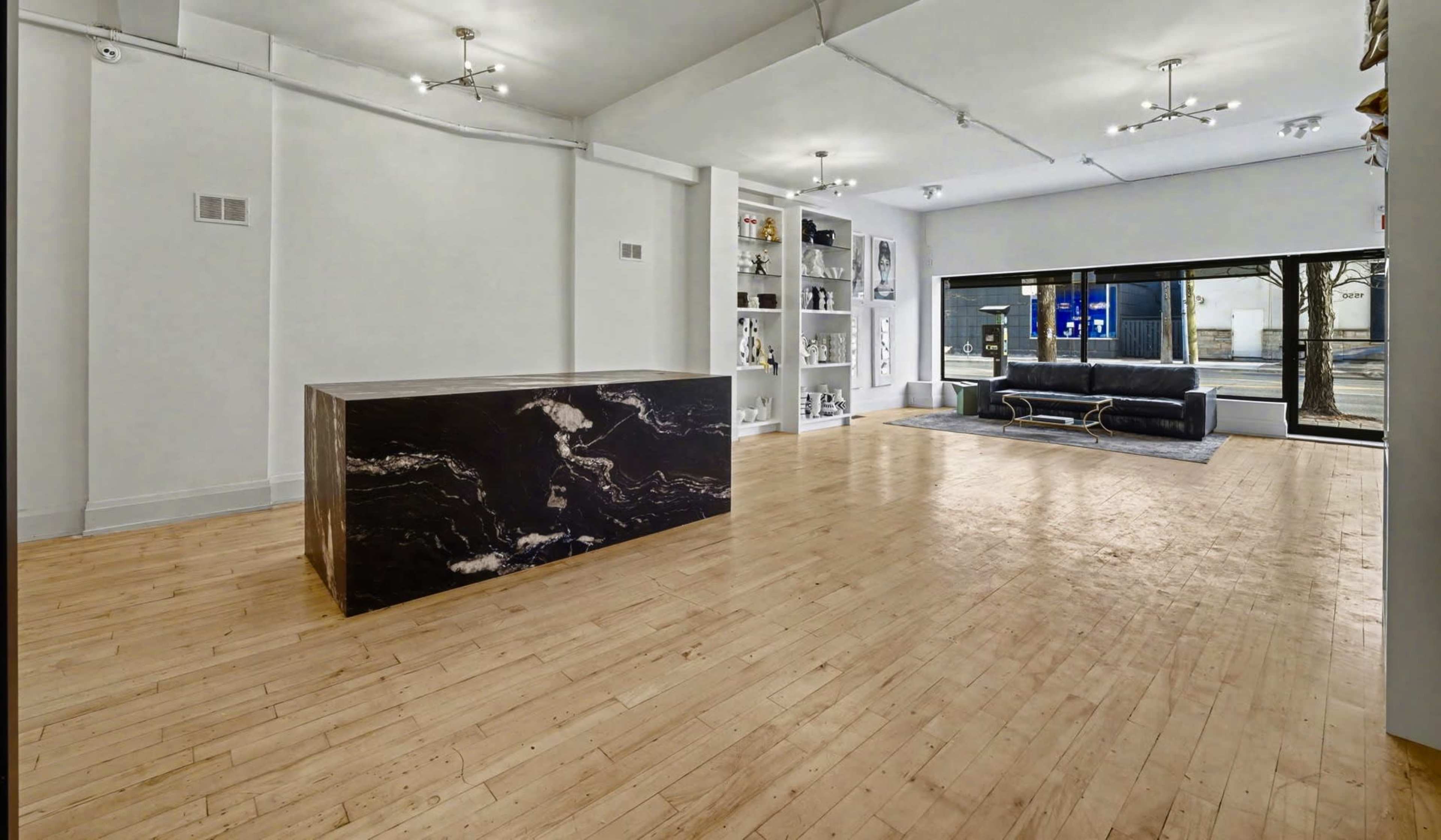 The image shows a spacious interior with a wooden floor, a large dark marble reception desk, shelves displaying various items, and a modern couch situated near large windows.