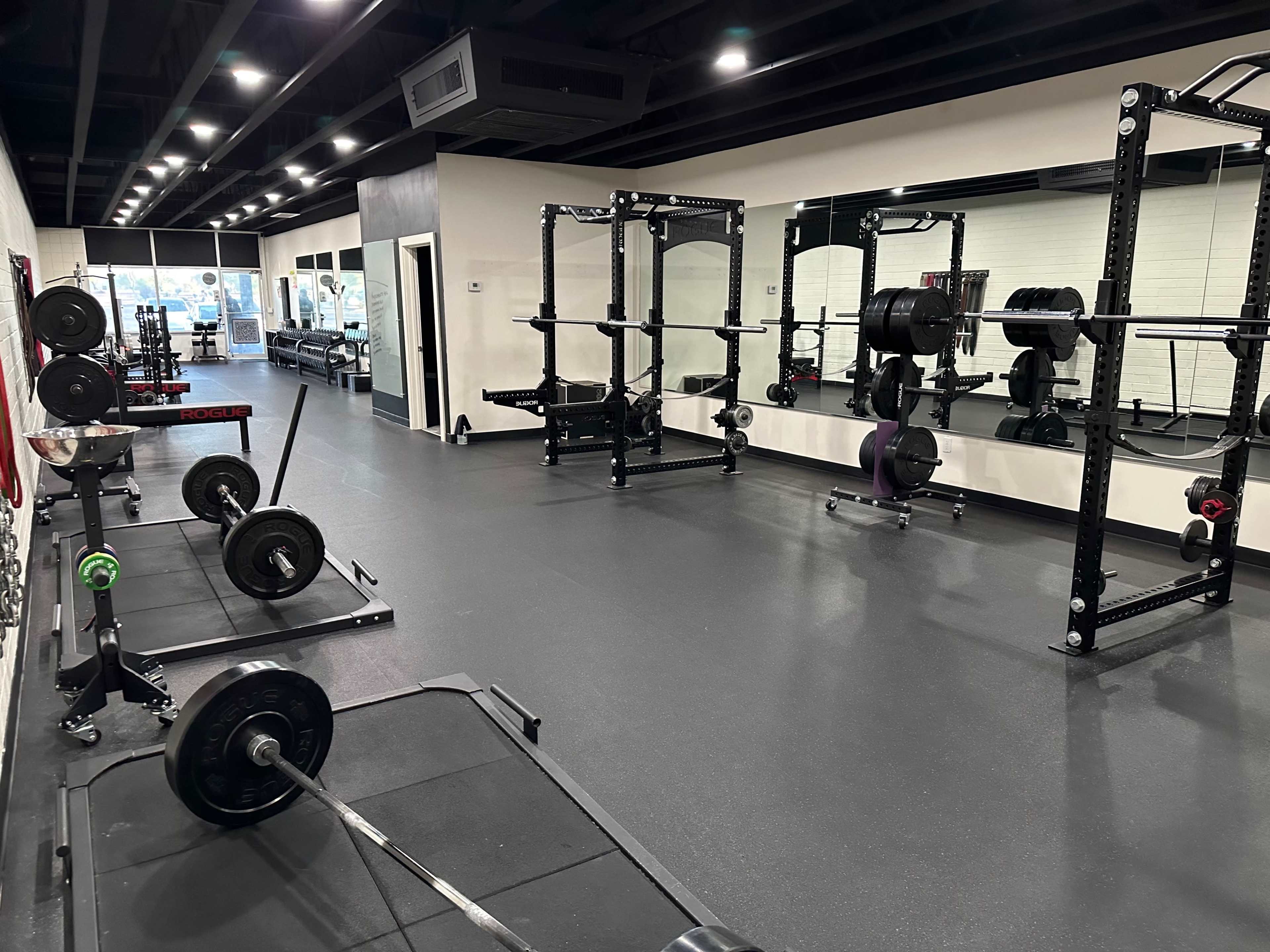 The image shows a modern gym interior with weightlifting equipment, including barbells and squat racks, set on a rubberized floor.