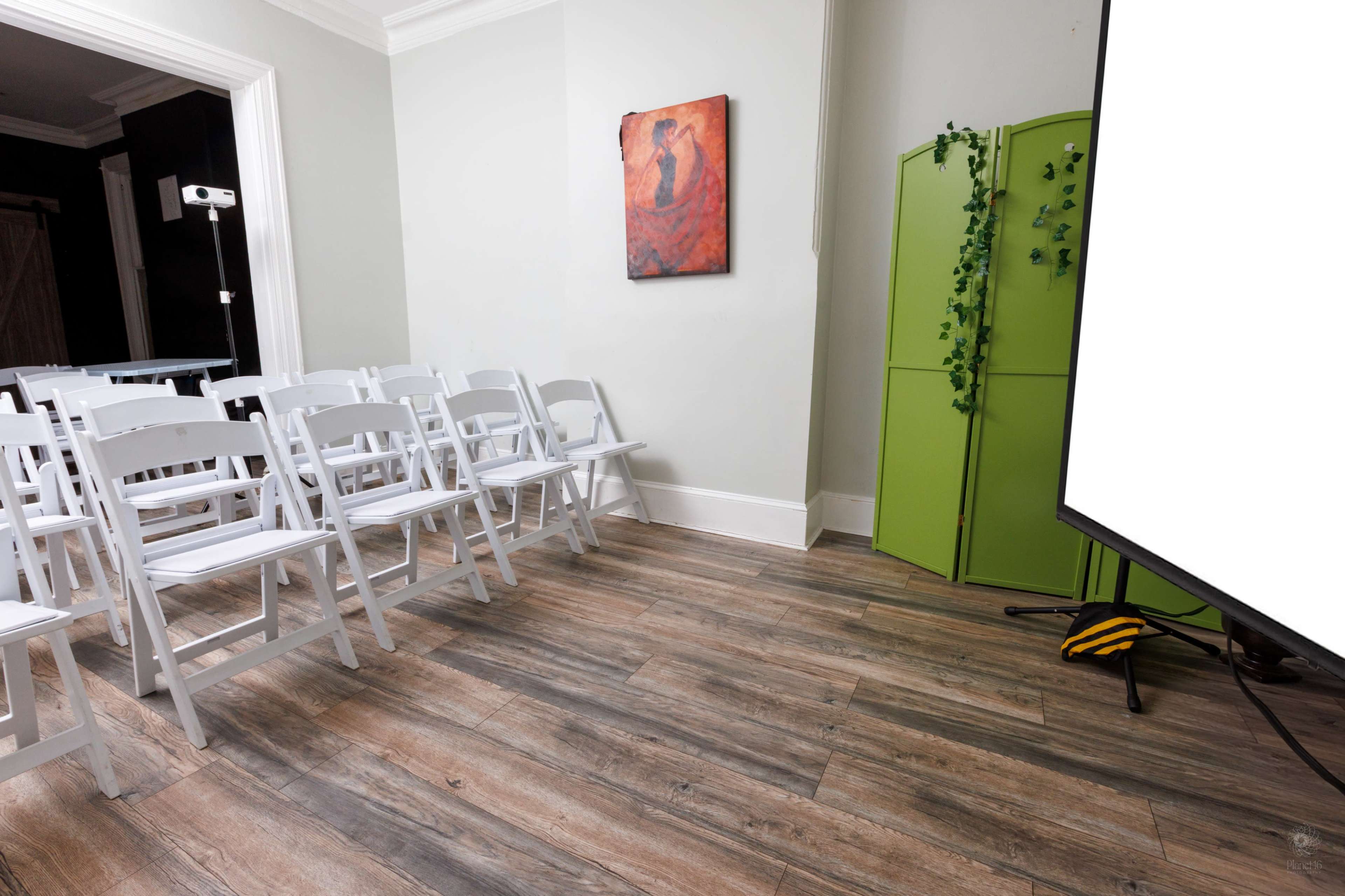 The image shows a room with white folding chairs arranged in rows, a green folding screen, and a large white projector screen on the right.