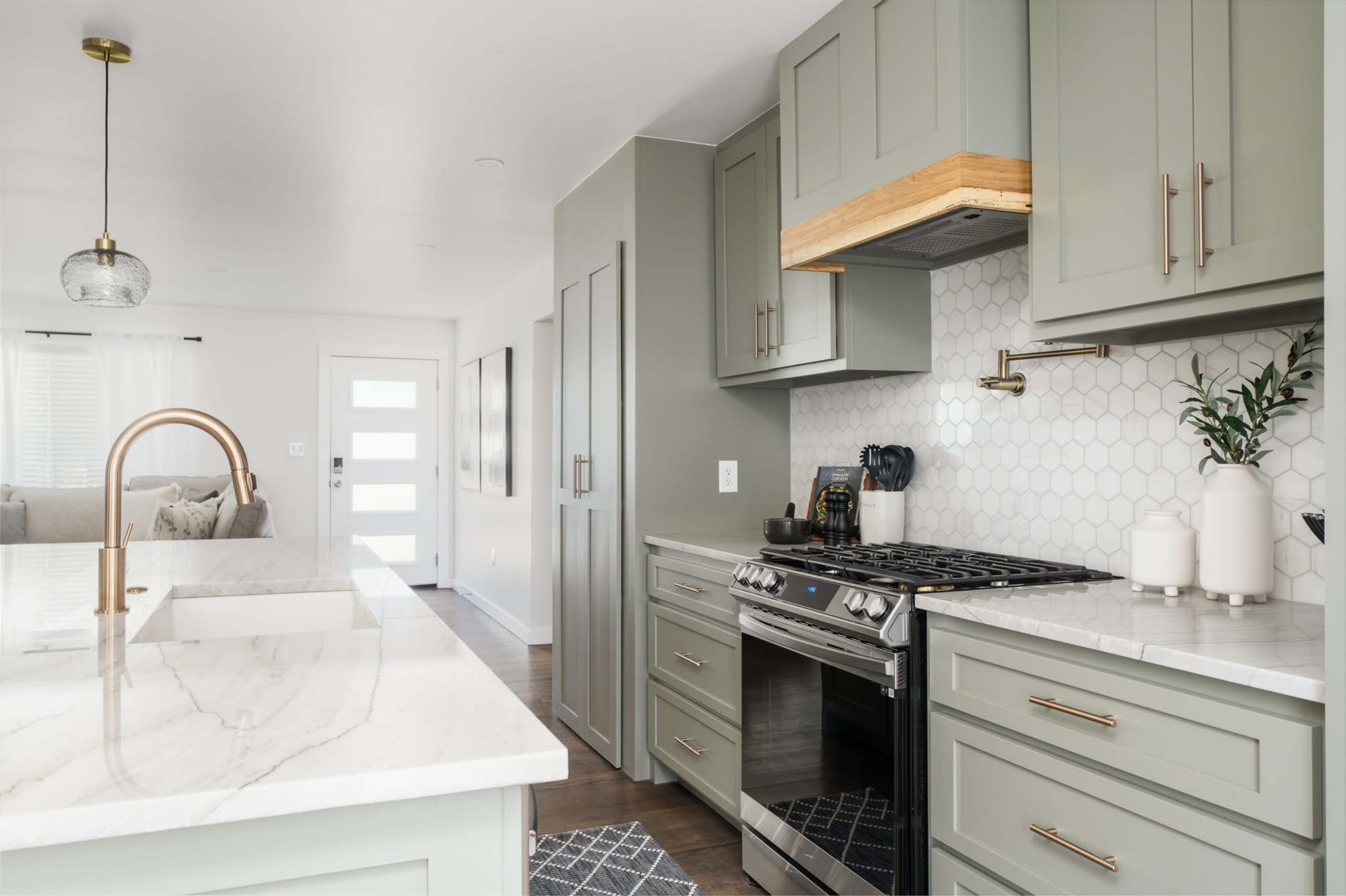 A modern kitchen features green cabinetry, a marble countertop, and a hexagonal tile backsplash with a gas stove and a wooden hood.