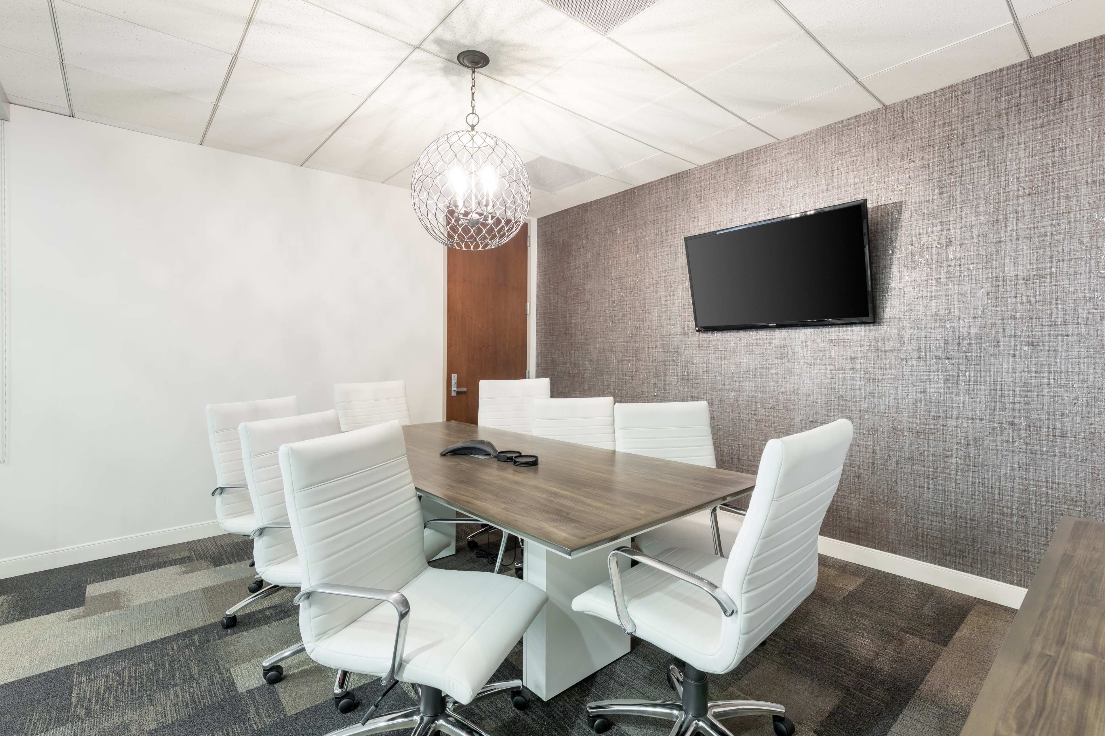 A modern conference room features a large wooden table surrounded by white ergonomic chairs, a wall-mounted television, and a decorative light fixture.