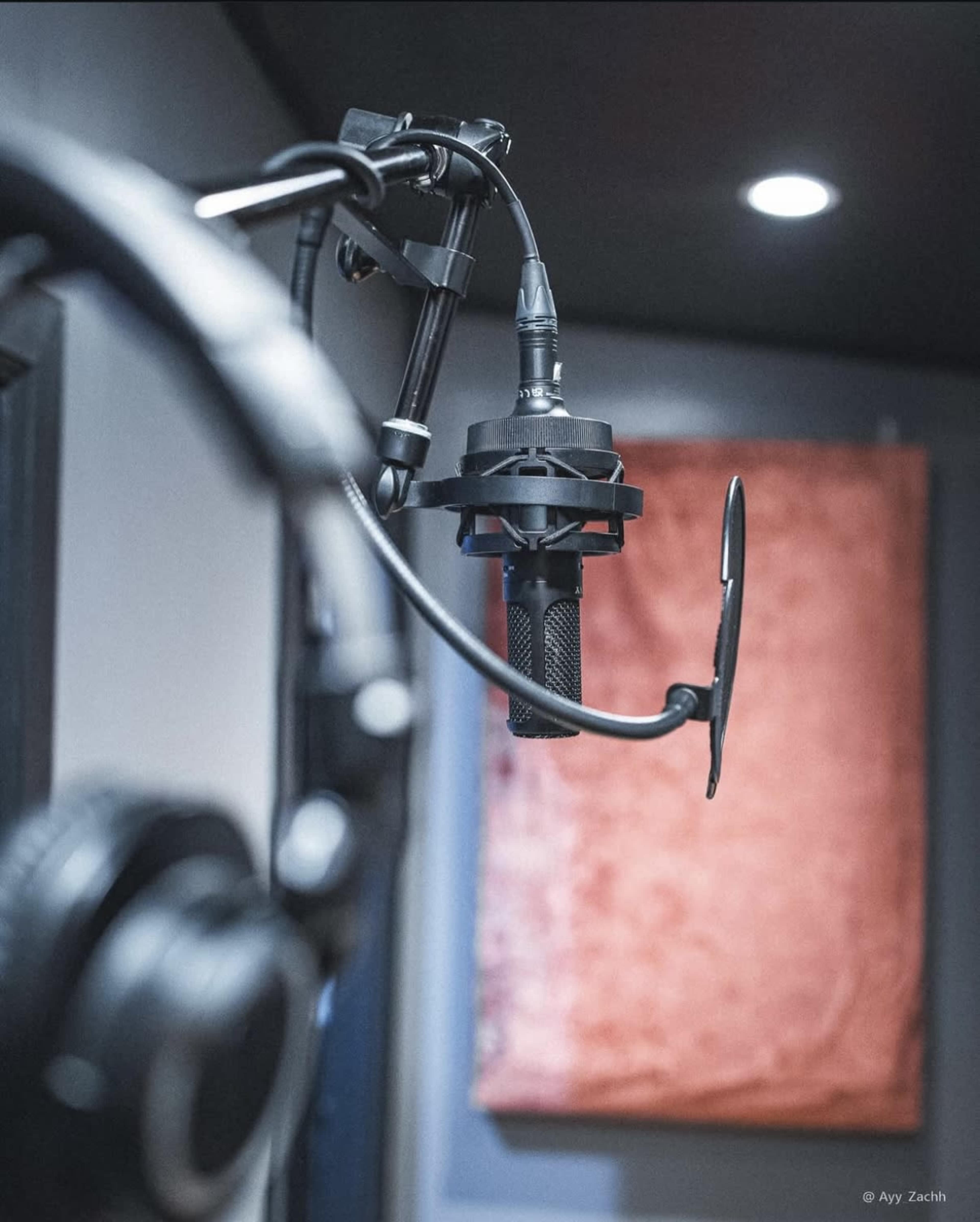 A microphone is mounted on a stand in a recording studio, with headphones hanging nearby and a textured wall in the background.