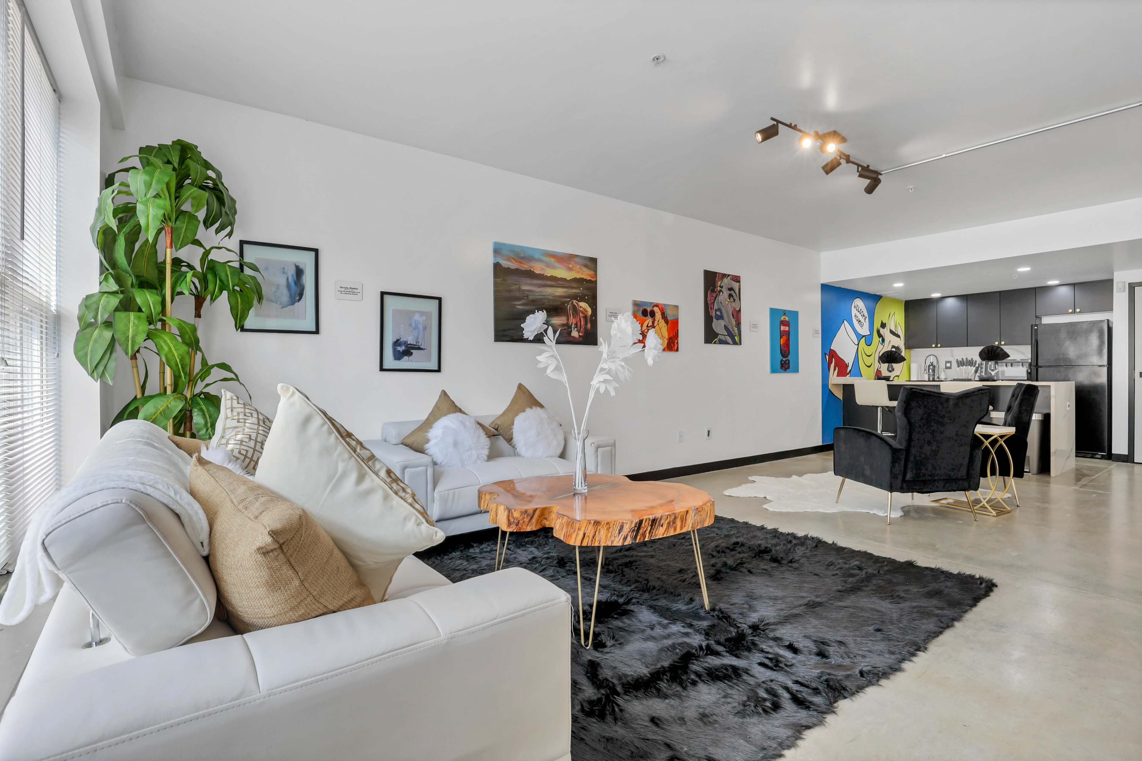 A modern living area featuring a white sofa, a coffee table with a natural wood finish, a black area rug, and colorful artwork on the walls, with a kitchen visible in the background.