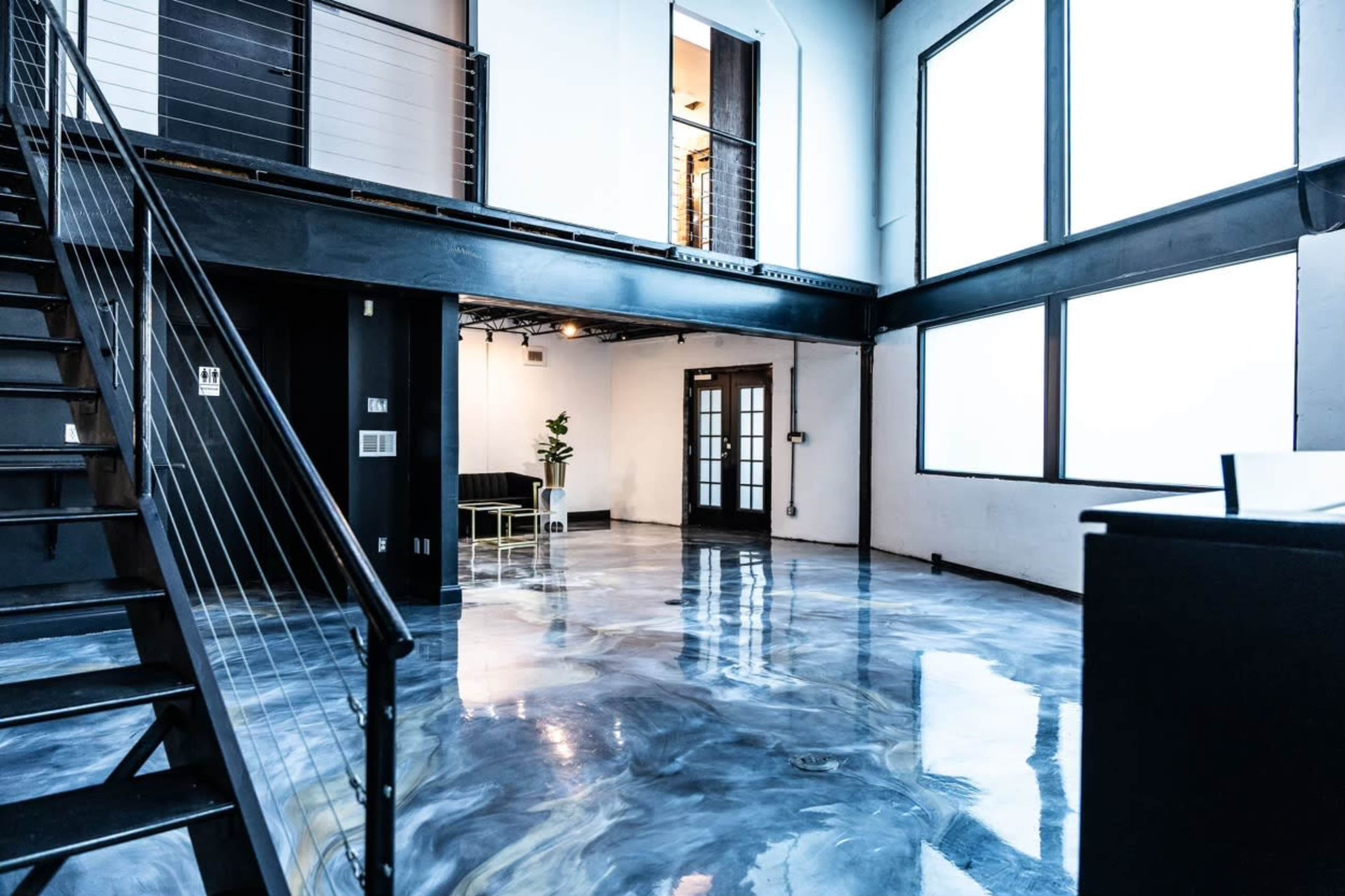 The image shows a modern, open interior space with polished concrete floors, large windows, and a staircase leading to an upper level.