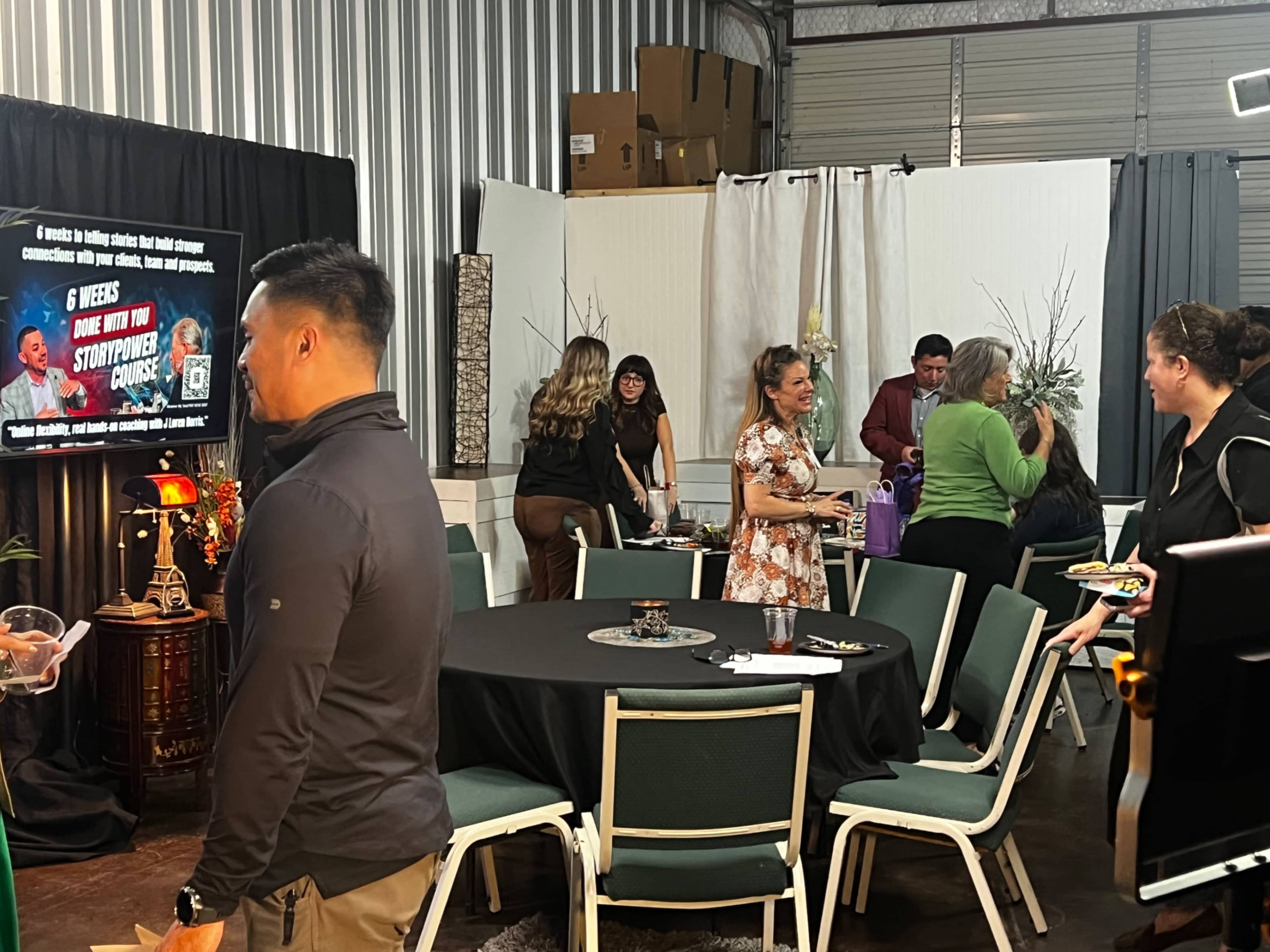 A group of people gathers in a decorated indoor space, with tables set for an event and a presentation screen in the background.