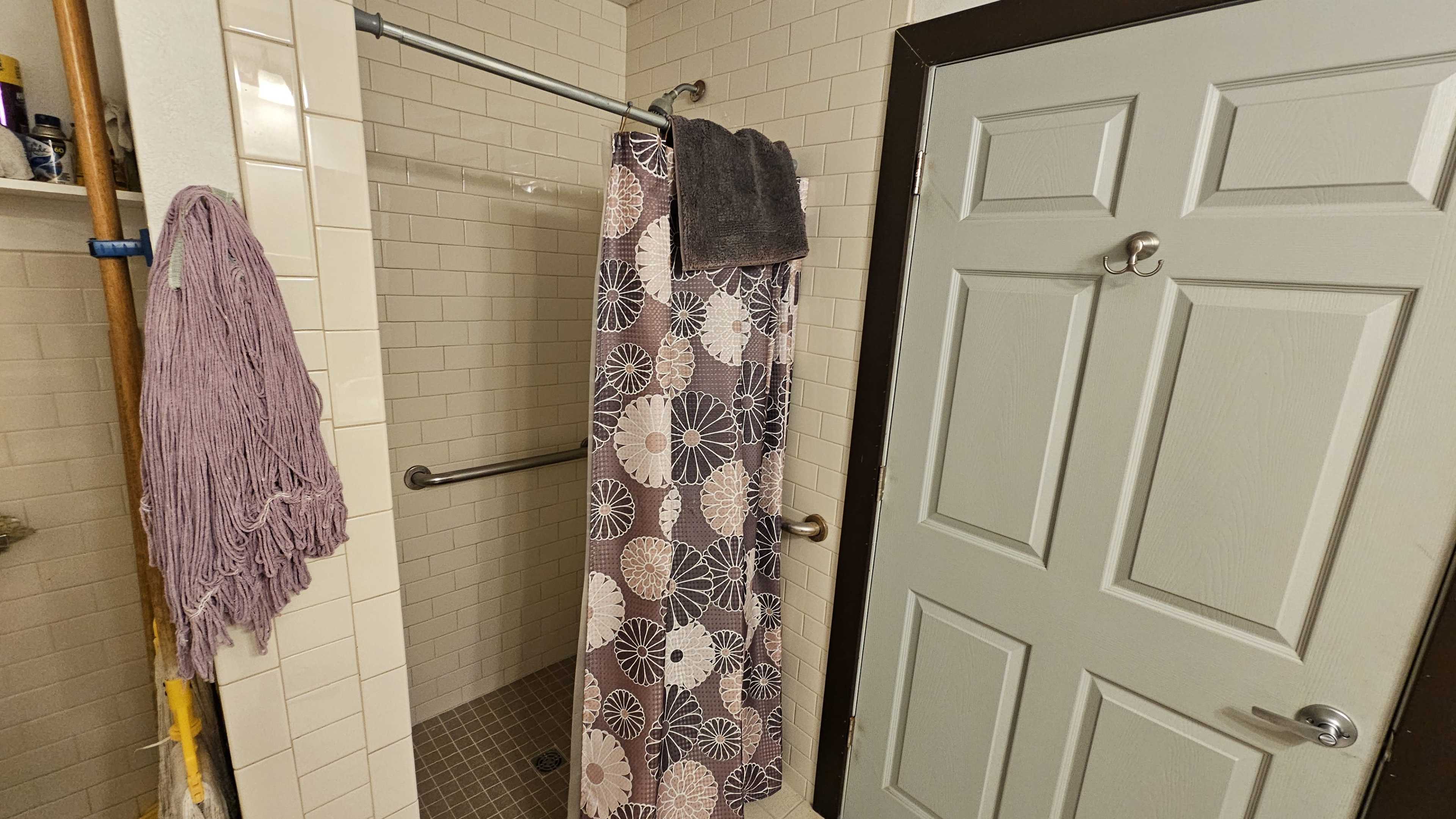 The image shows a bathroom with a shower area, a floral-patterned shower curtain hanging, and a light-colored door.