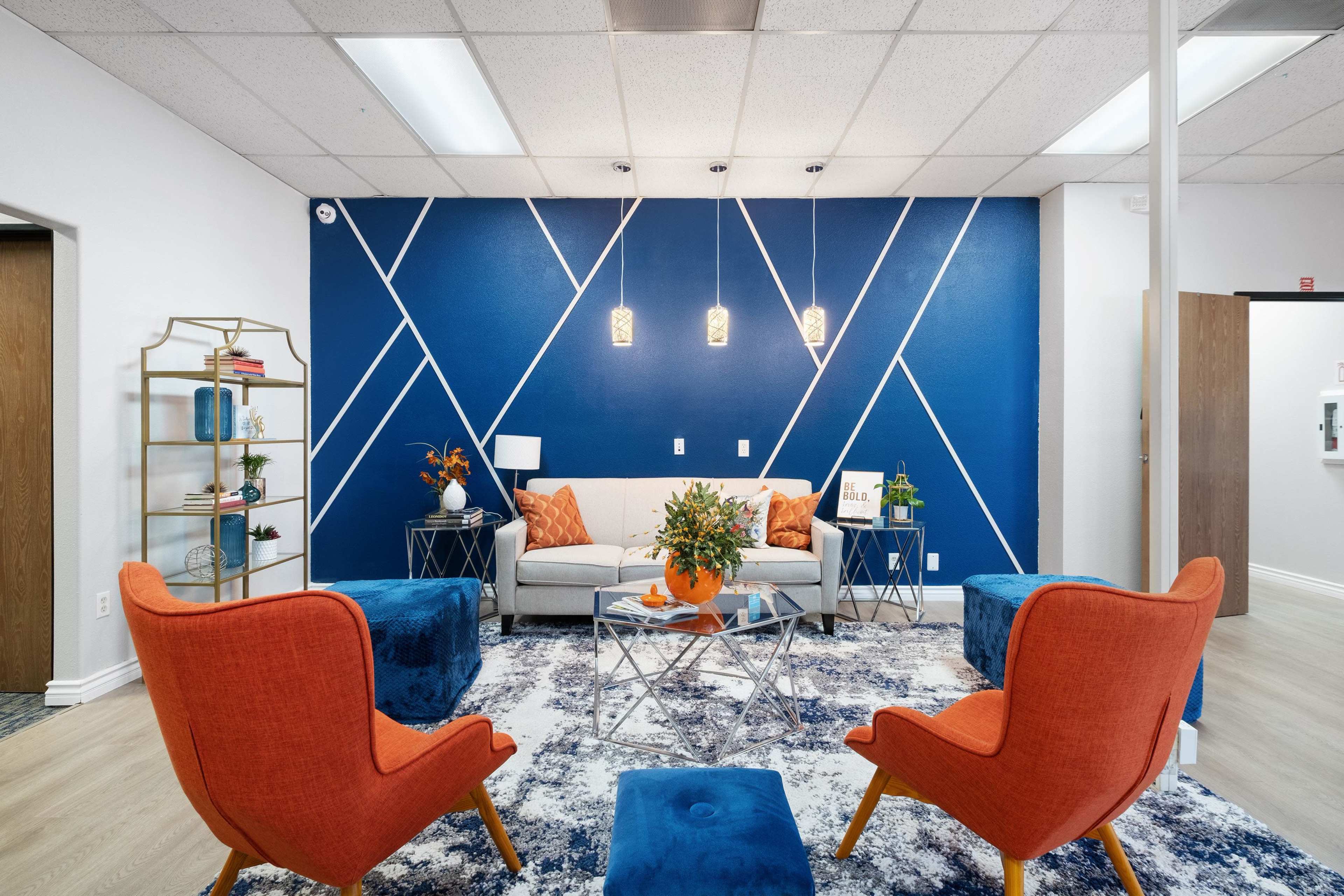 A modern seating area with orange chairs, a gray sofa, and a blue geometric accent wall, complemented by a decorative rug and plants.