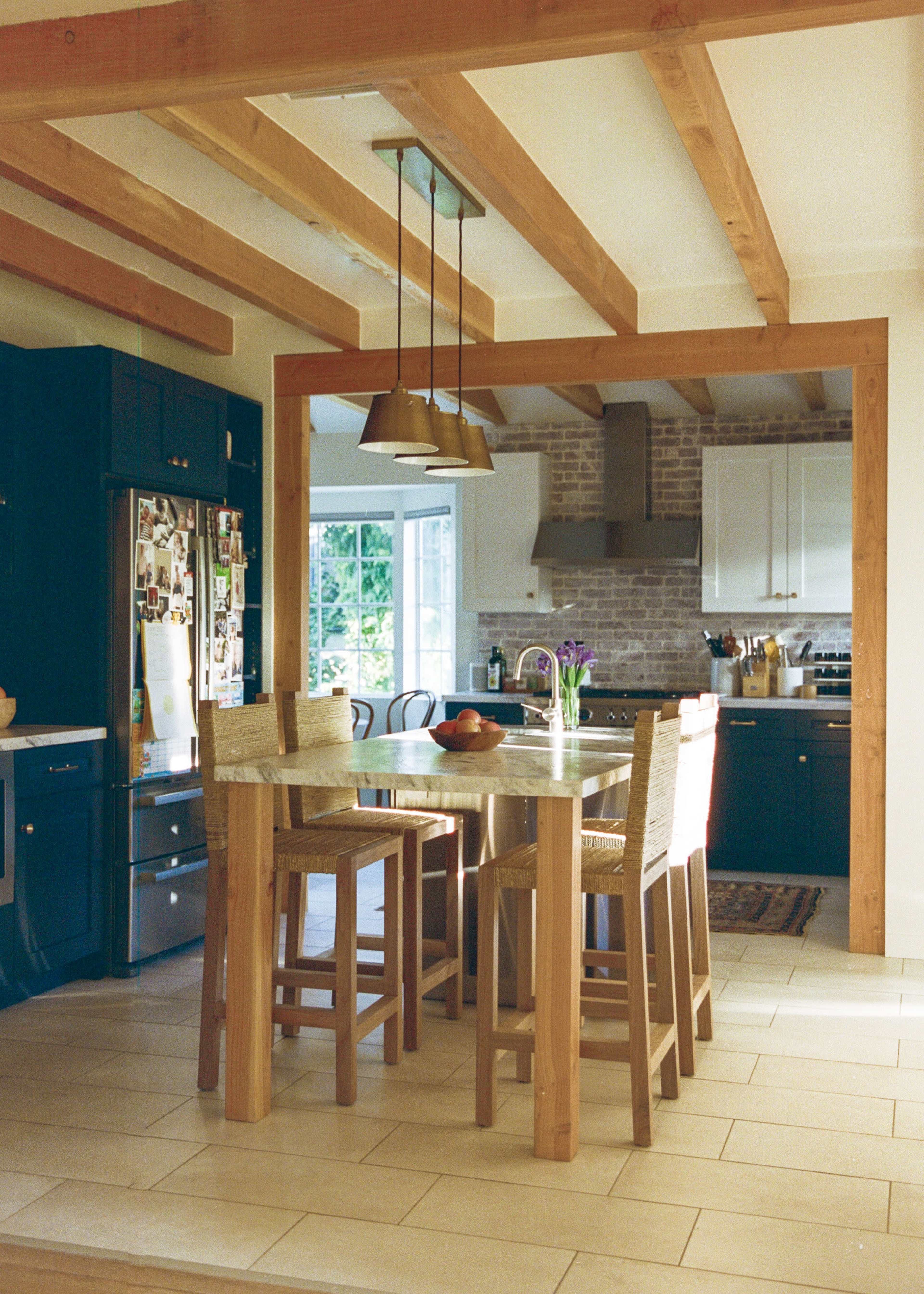 A kitchen with exposed wooden beams features a dining table surrounded by four wooden chairs, with cabinets and a brick backsplash in the background.