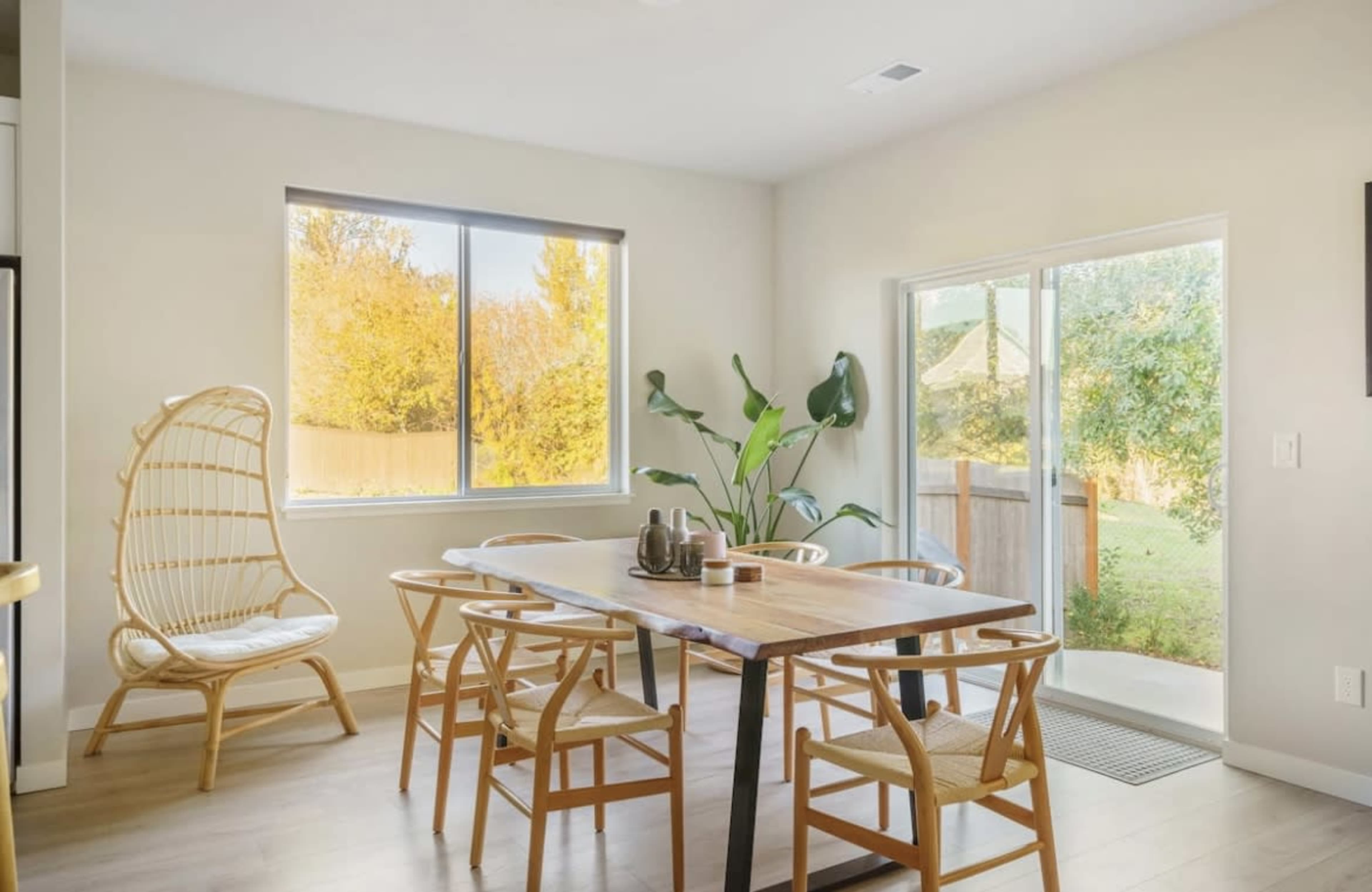 A dining area features a wooden table surrounded by chairs, with a large window and sliding door revealing a view of a backyard.