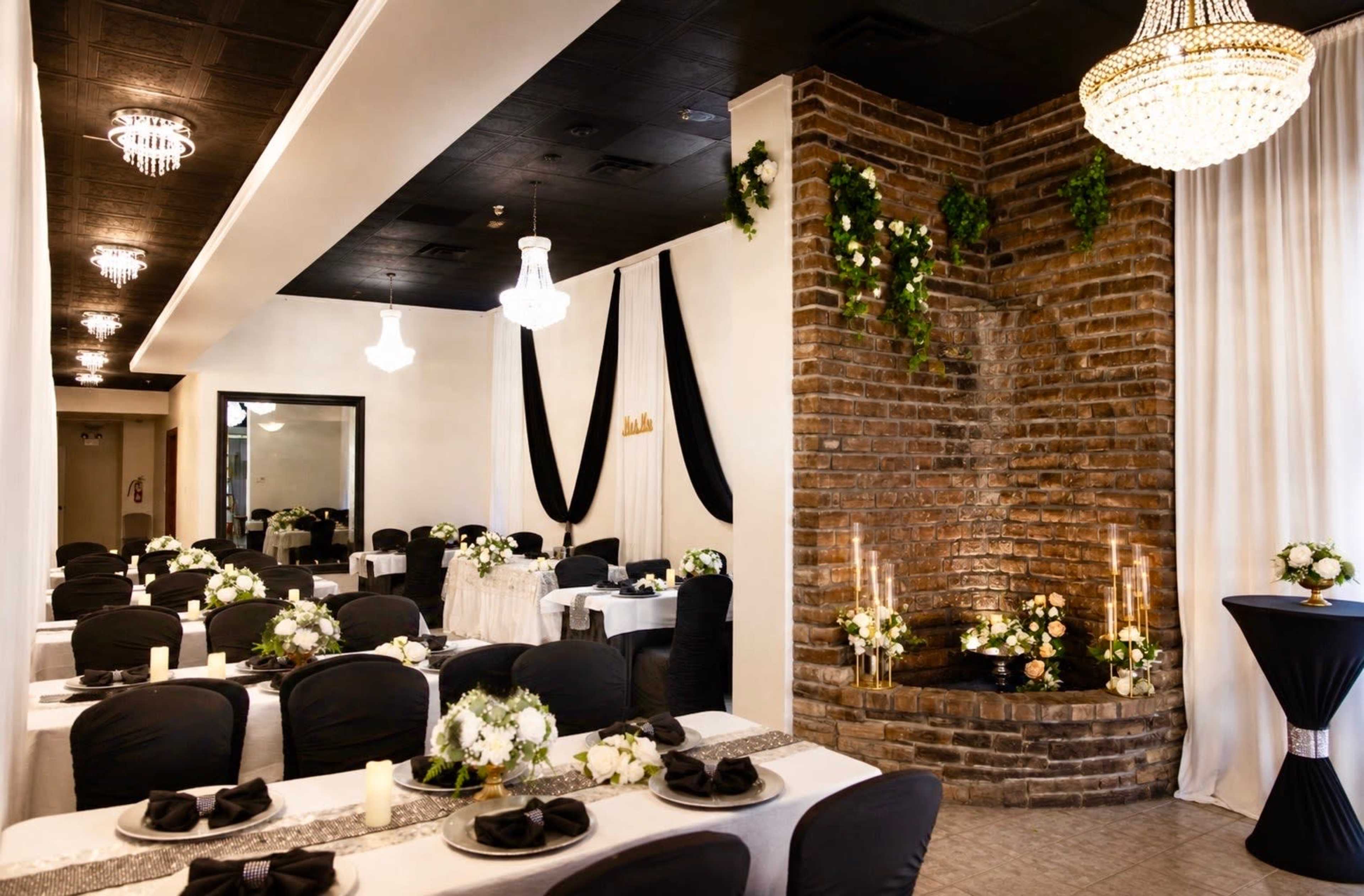 A dining area features elegantly arranged tables with black and white settings, complemented by a brick fireplace adorned with greenery and flowers.