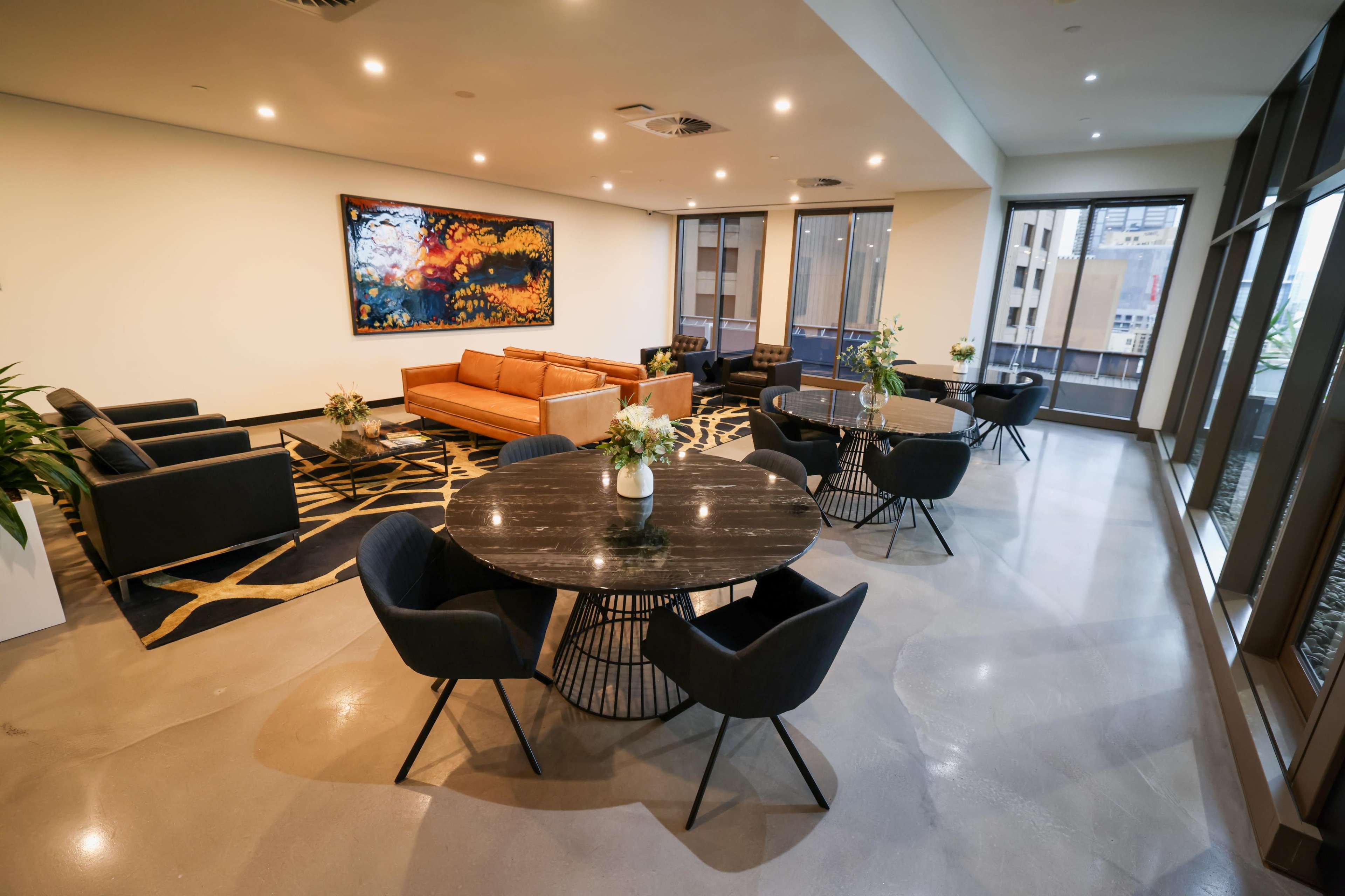 The image shows a modern lounge area with seating arrangements, including a bright orange sofa and dark-colored chairs, complemented by round tables and large windows.