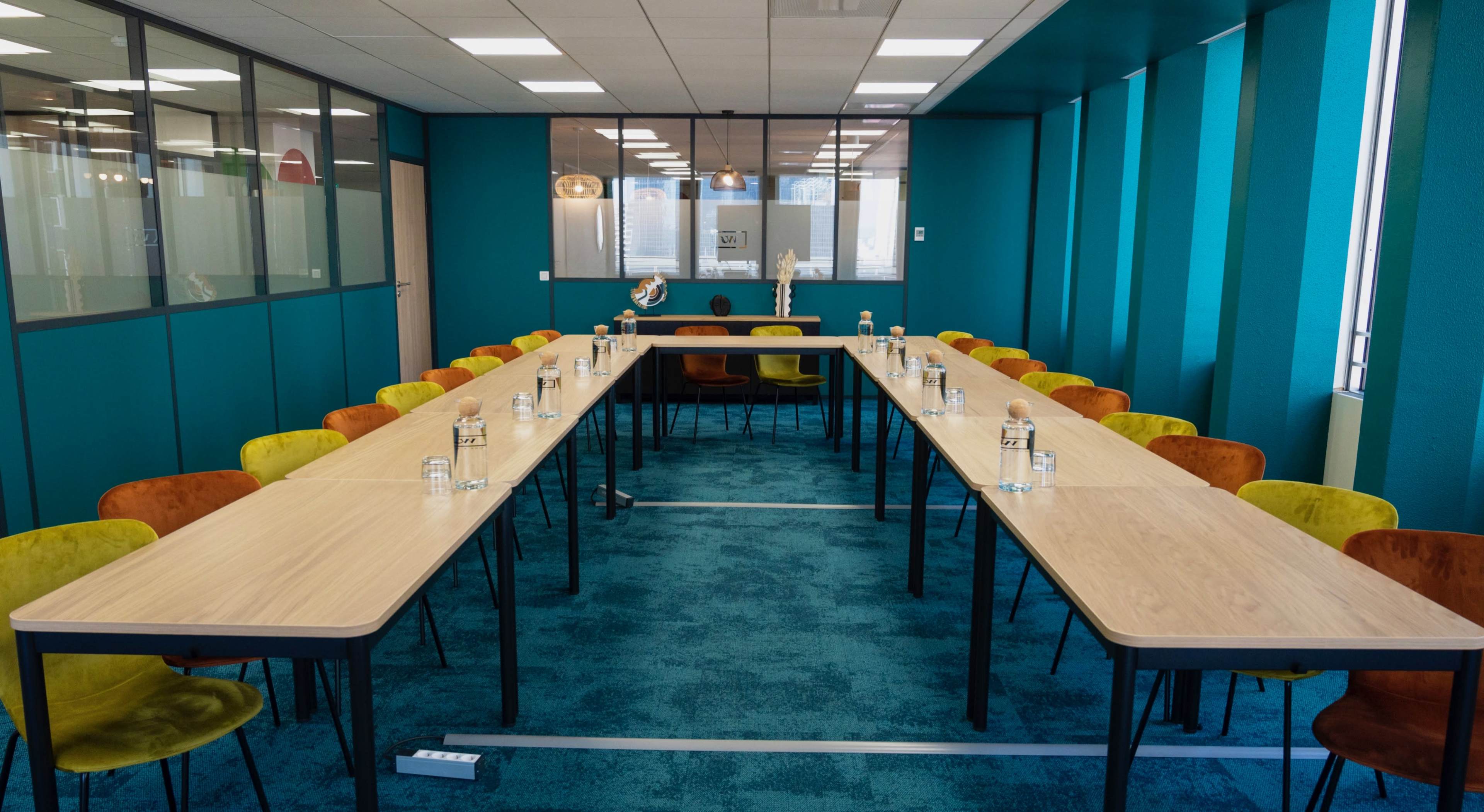 A conference room featuring two long tables arranged in a U-shape, surrounded by colorful chairs and illuminated by natural light.