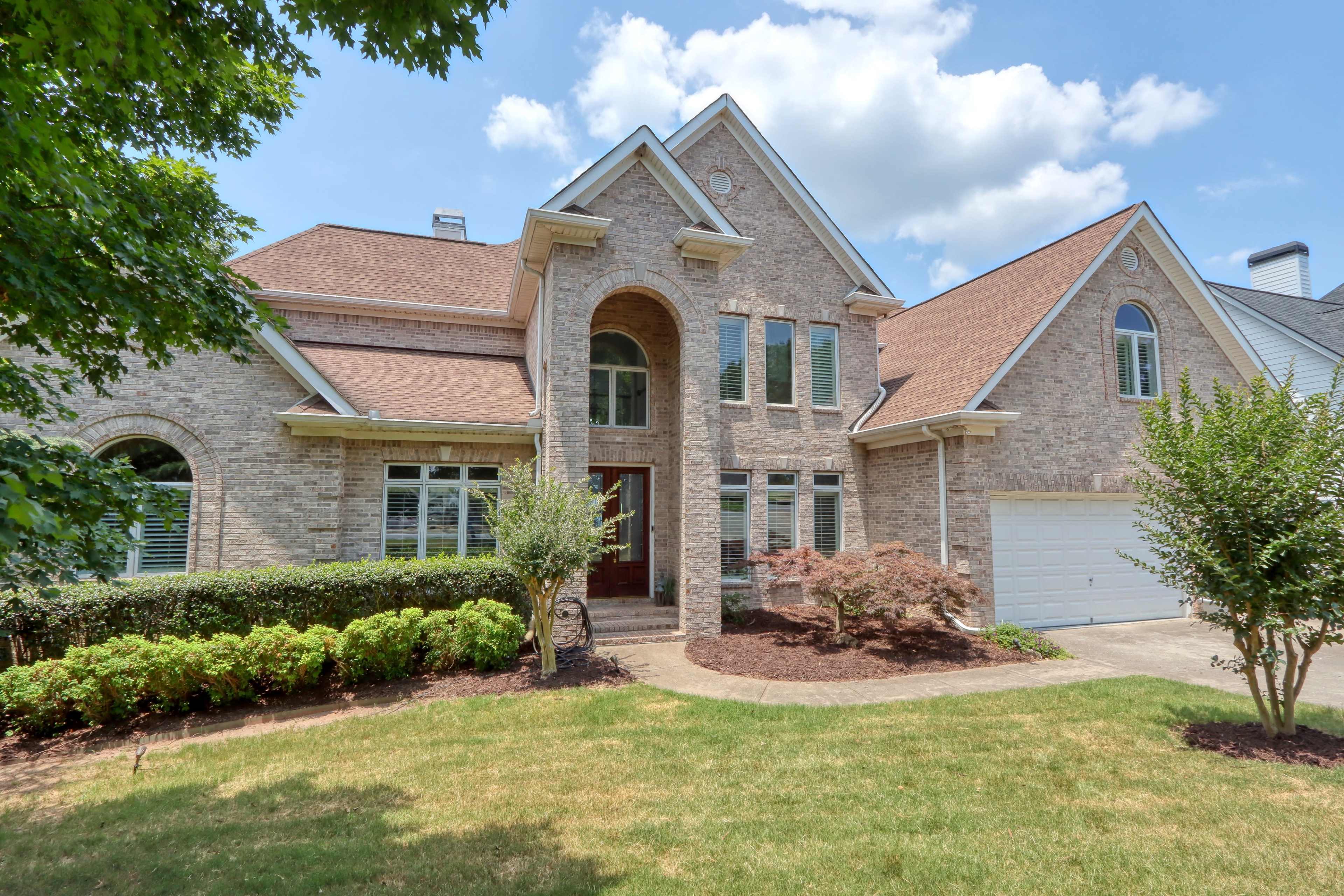 A large brick house with a peaked roof, multiple windows, and a two-car garage is surrounded by neatly trimmed shrubs and a grassy lawn.