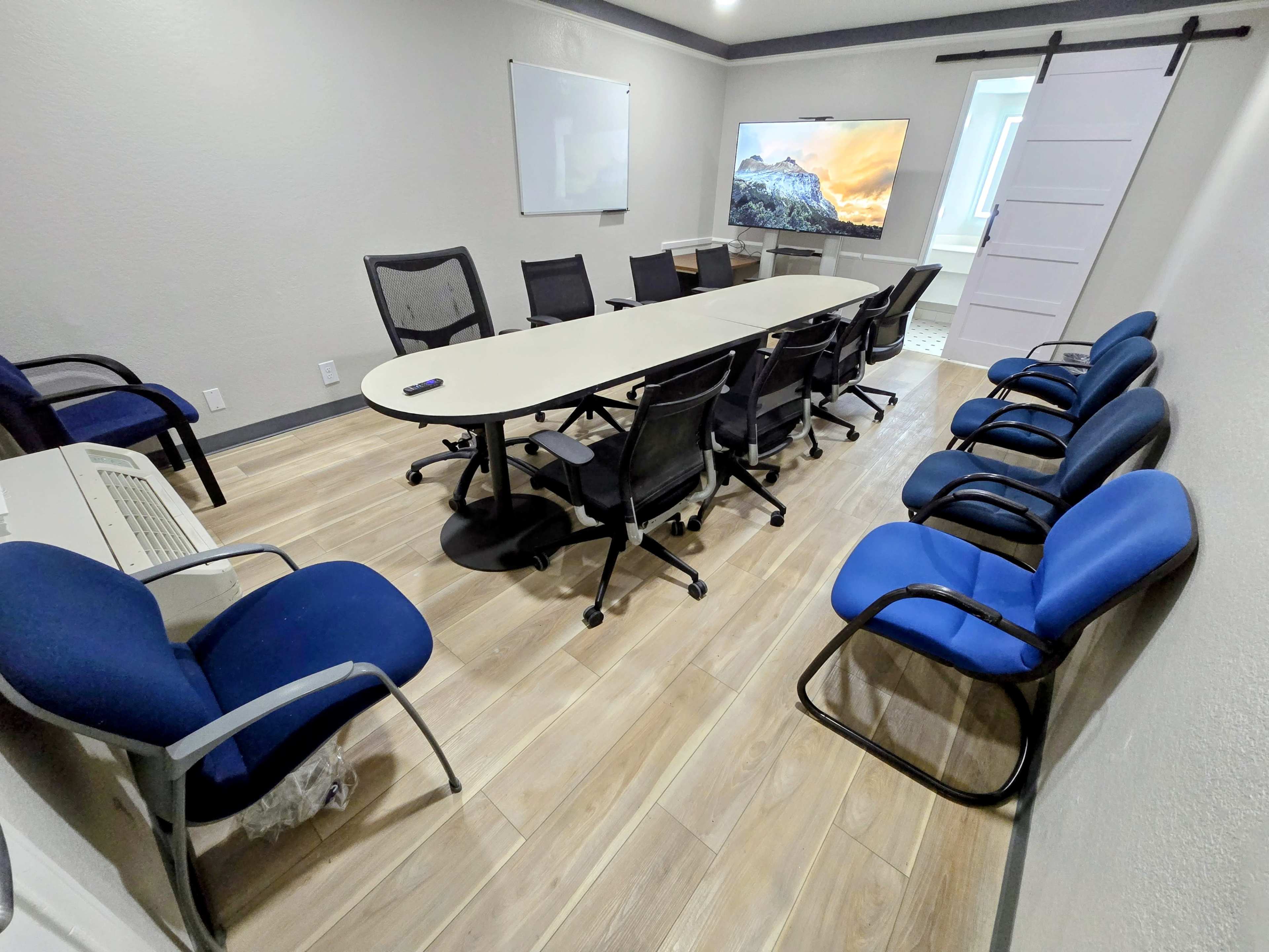 A conference room is equipped with a long table surrounded by black and blue chairs, featuring a large screen displaying a landscape image on one wall.