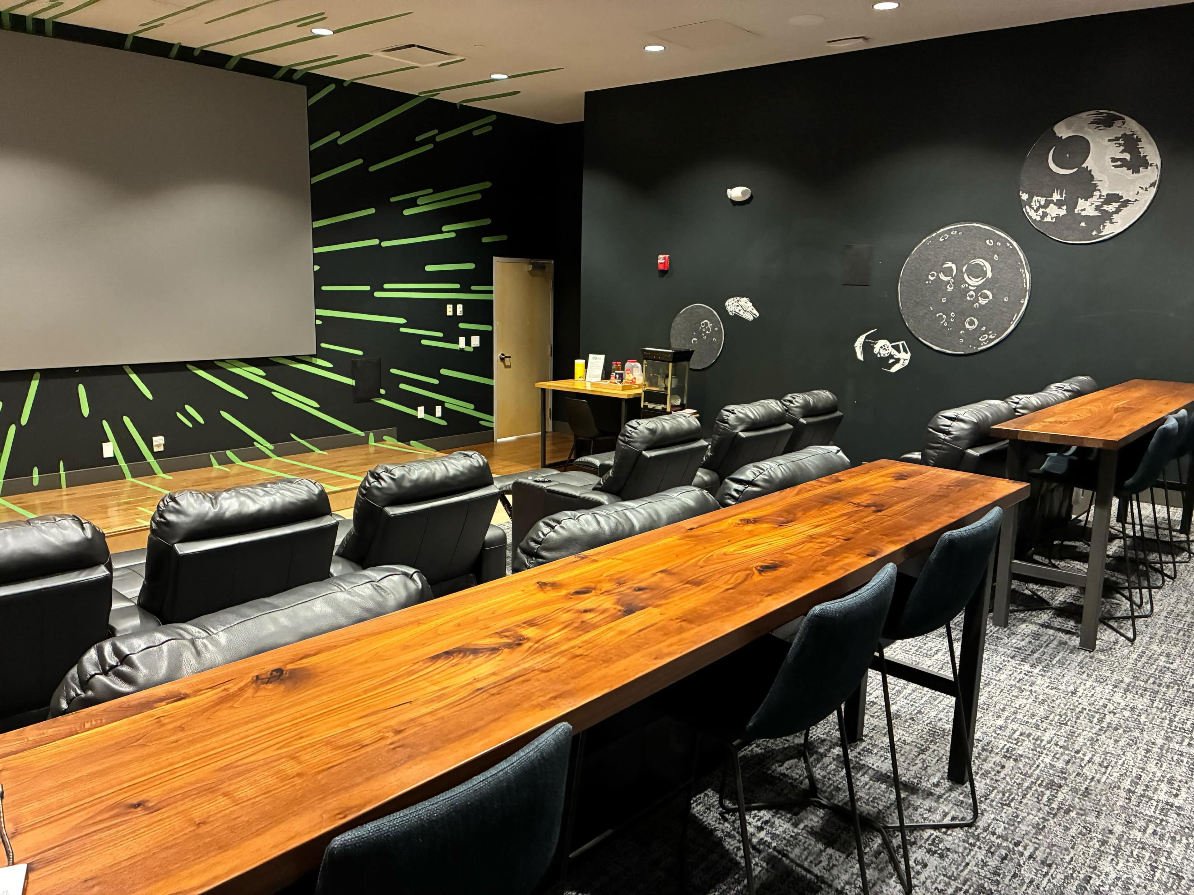 The image shows a modern presentation room with black leather seating, long wooden tables, and a projector screen on one wall.