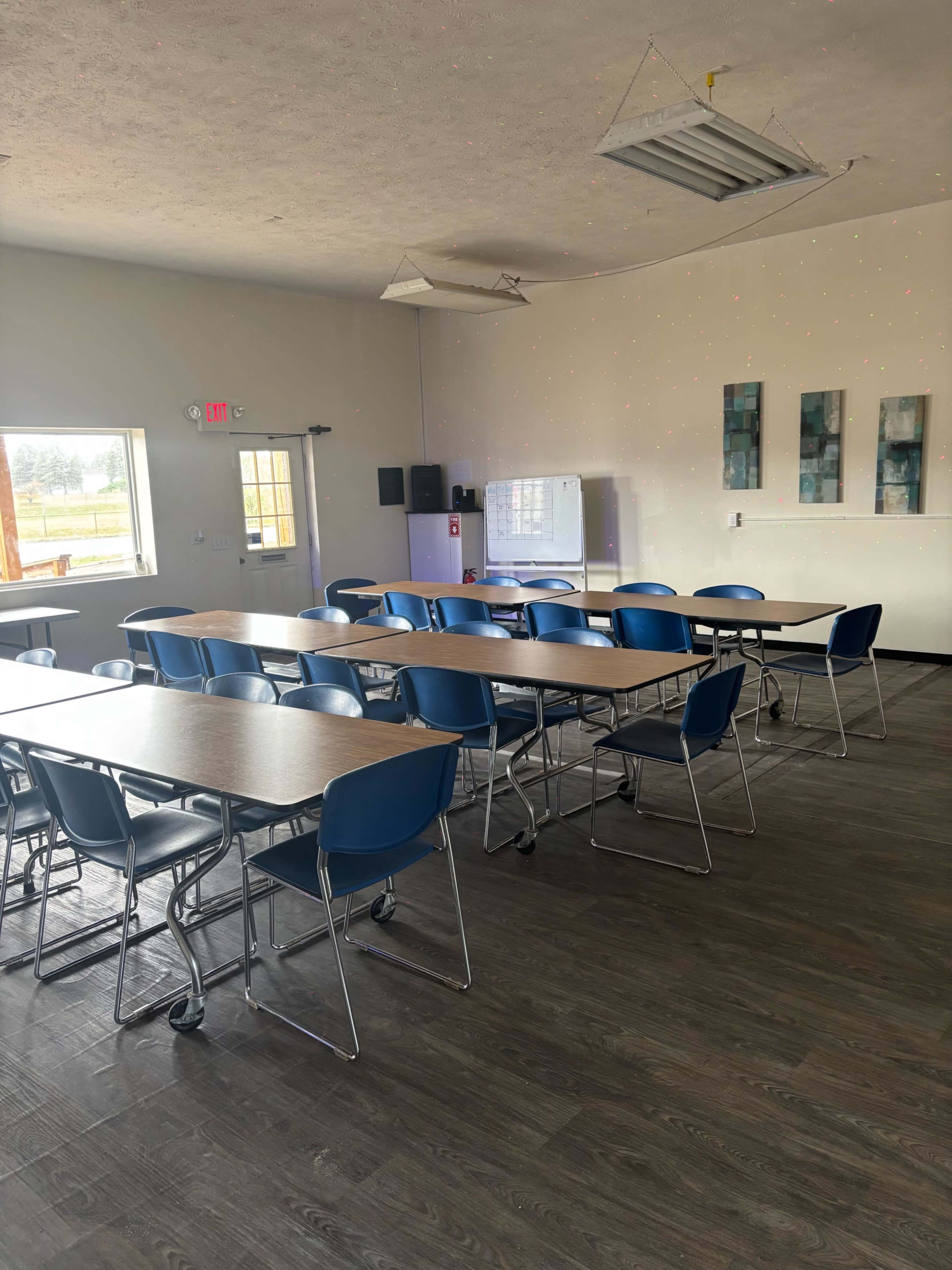 The image shows a spacious classroom with several rows of tables and blue chairs, along with a whiteboard and wall decorations in the background.