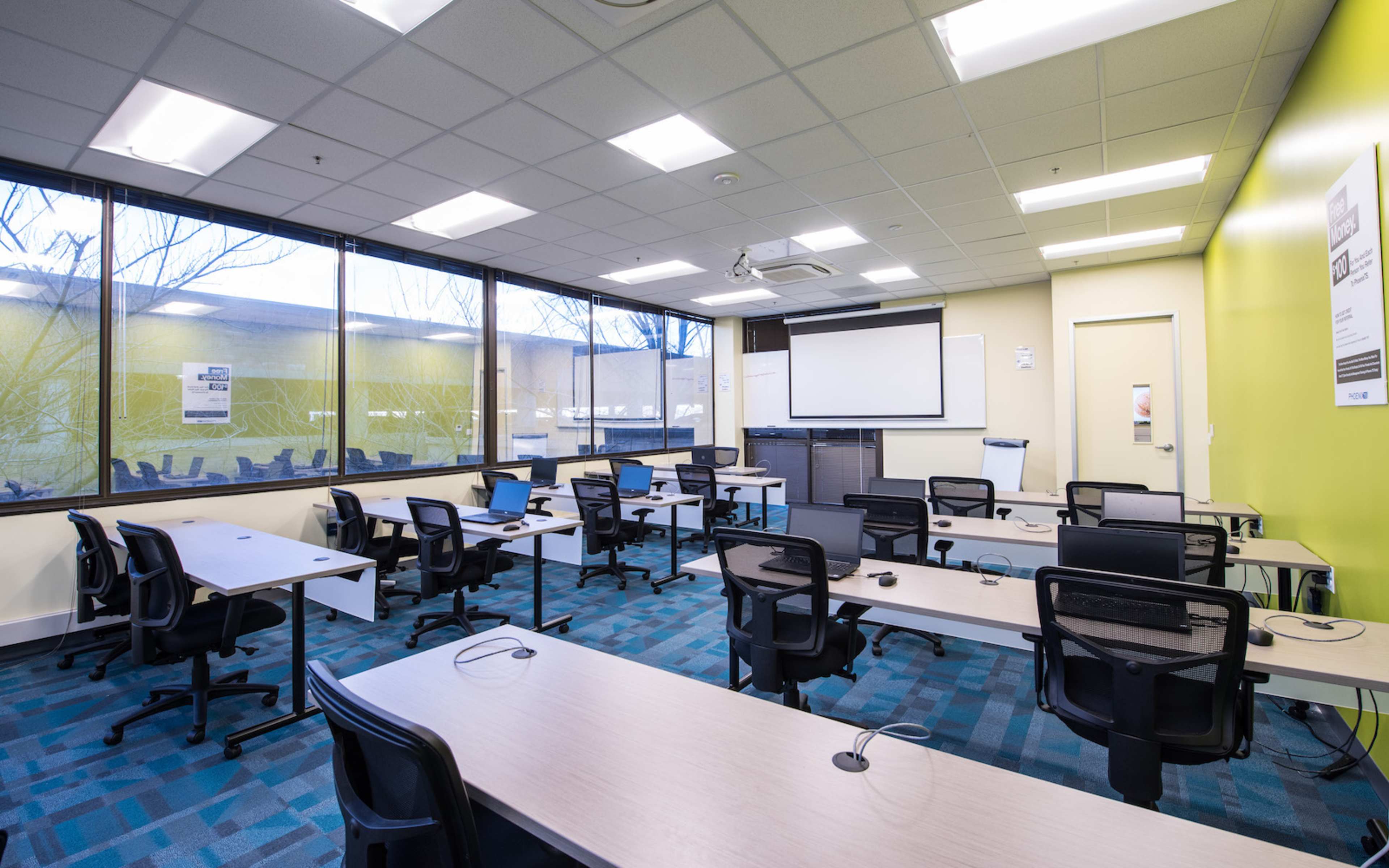 The image shows a modern classroom with multiple desks and chairs arranged in a spacious layout, large windows providing natural light, and a projector screen at the front of the room.