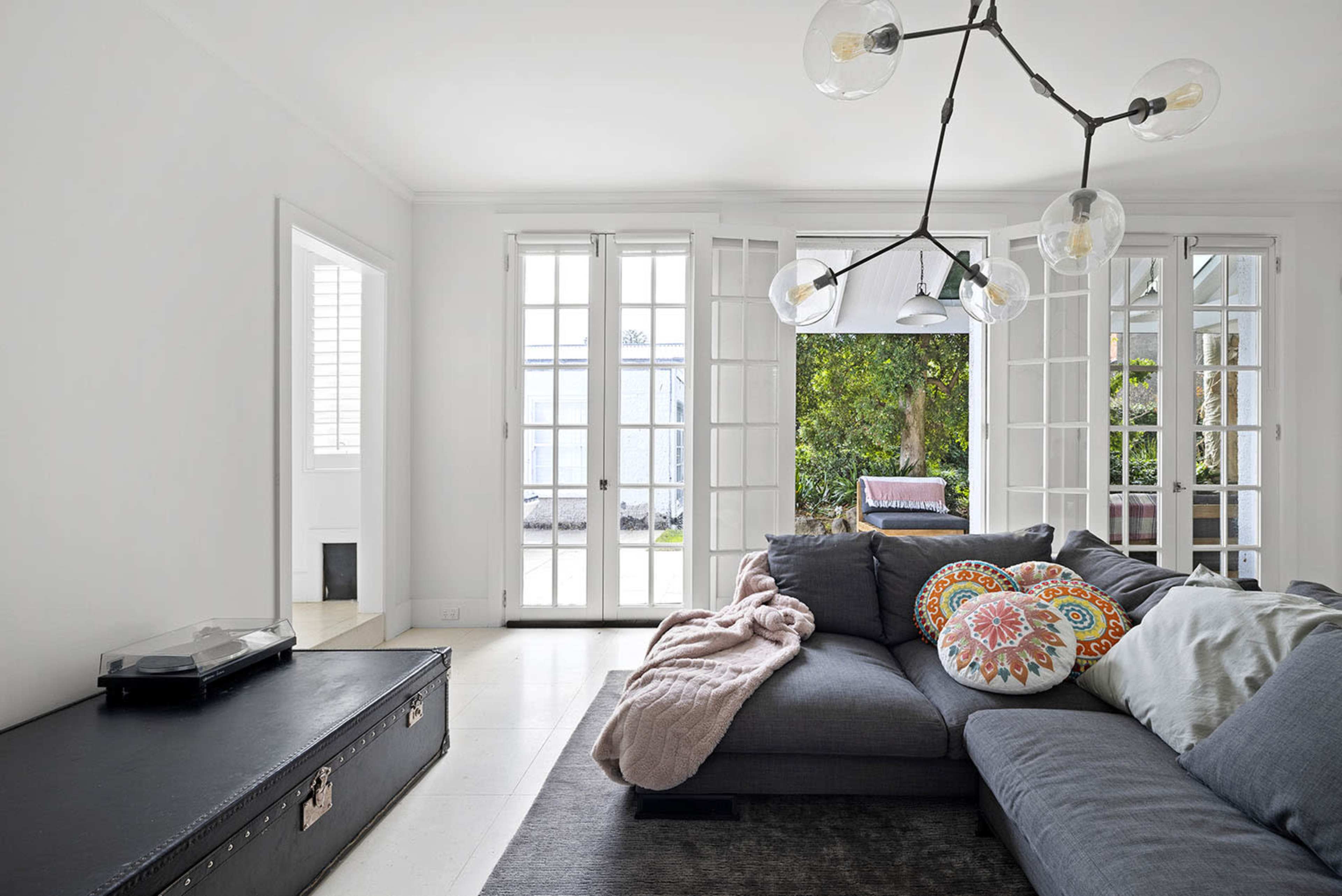 A modern living room features a gray sectional sofa with decorative cushions, a vintage suitcase-style coffee table, and large windows opening to a garden area.