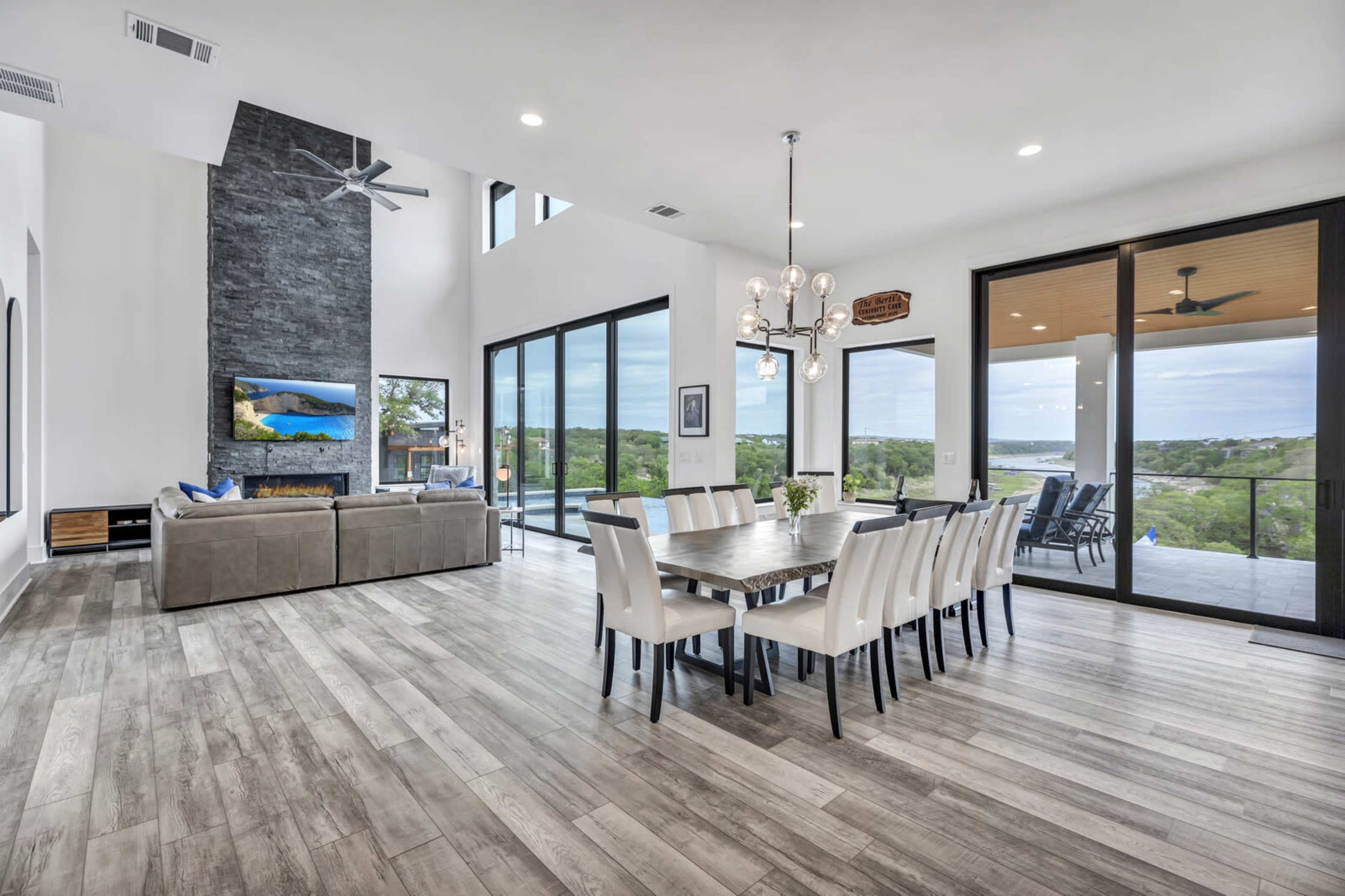 A modern, open-concept living and dining area with large windows overlooking a scenic landscape, featuring a long dining table and a sectional sofa.