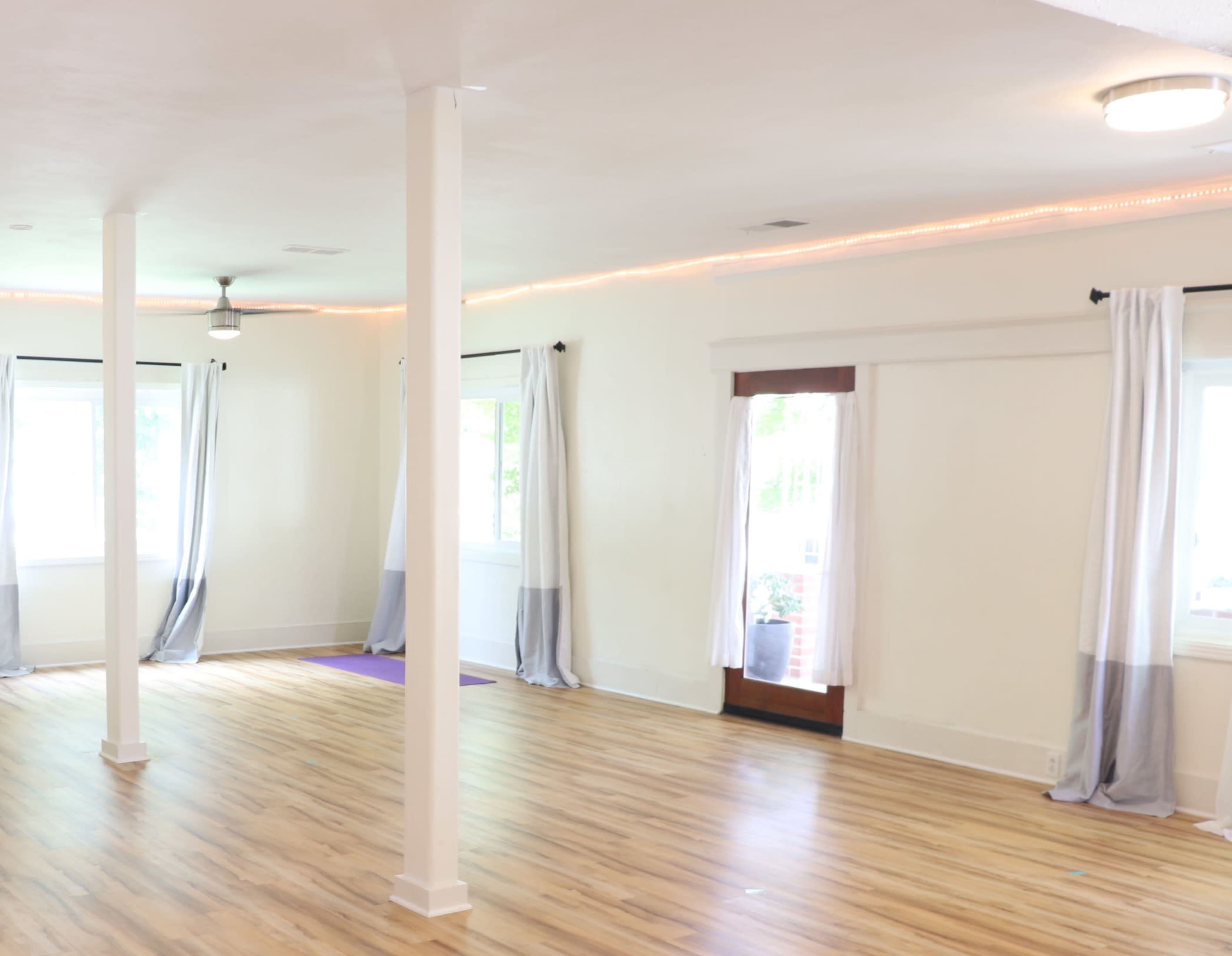 A spacious room with wooden floors, large windows, and white curtains featuring grey accents, along with two columns and a ceiling fan.