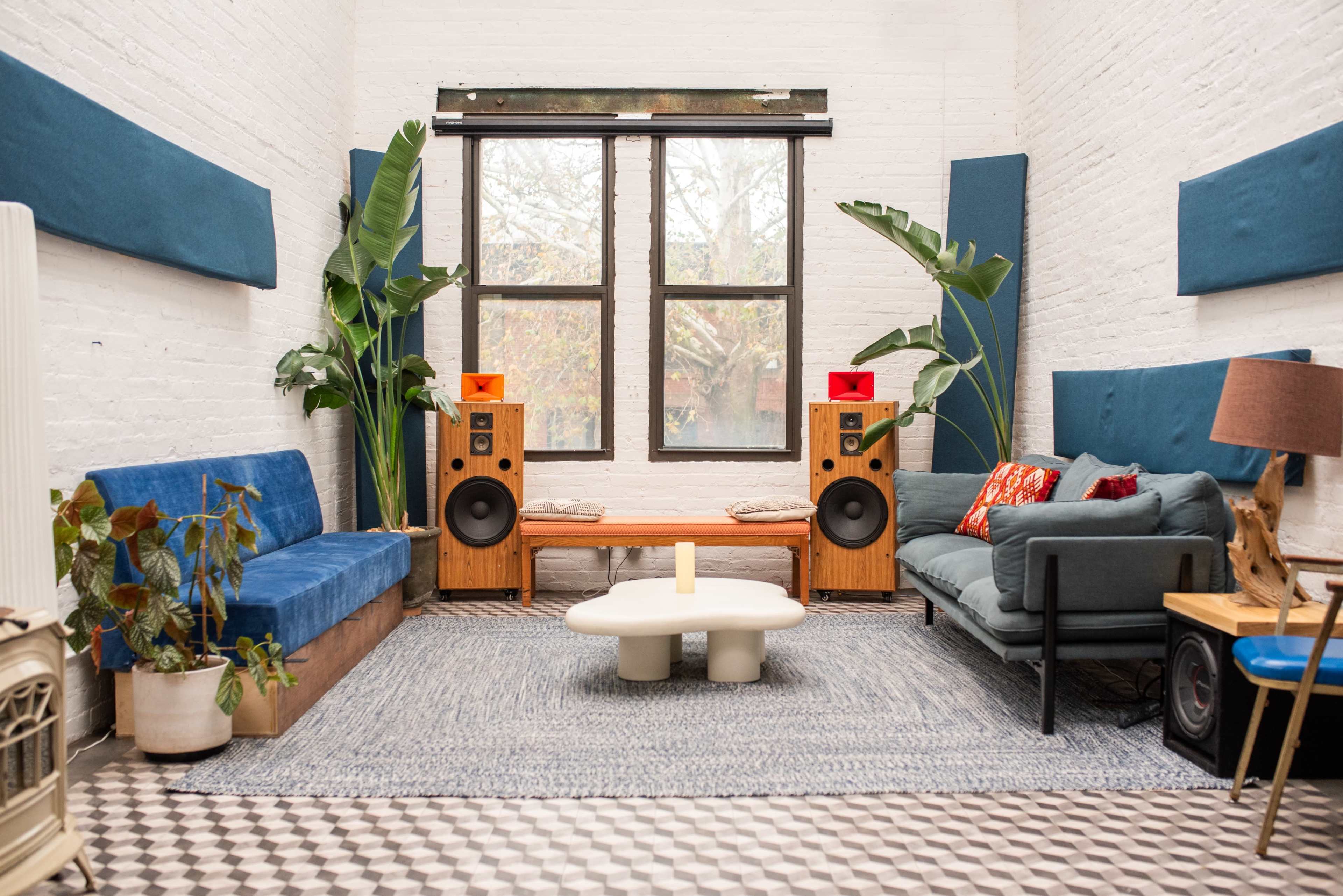The image shows a stylish living room with blue sofas, wooden speakers, a coffee table, and large windows allowing natural light in, accented by indoor plants.