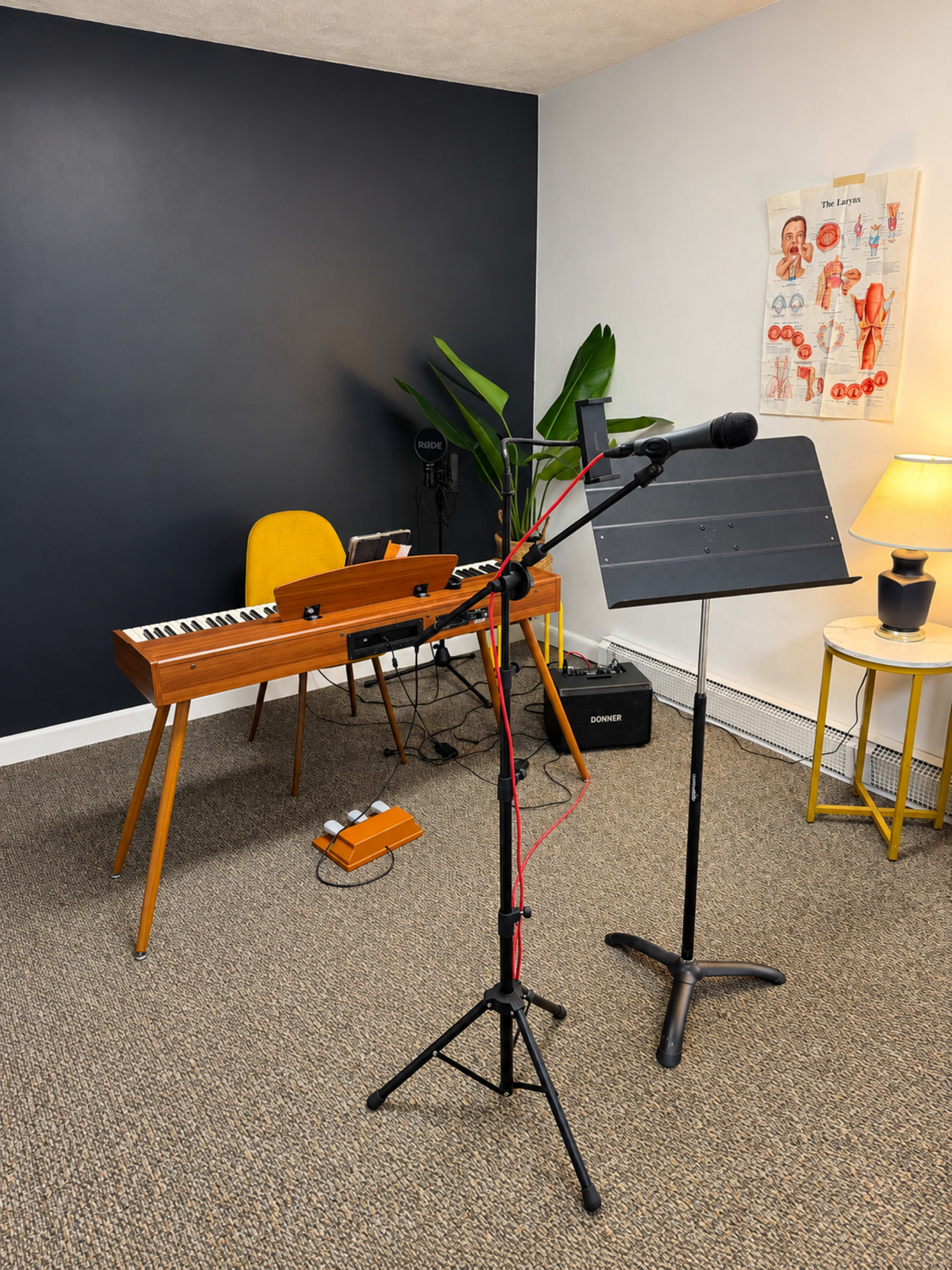 The image shows a small music studio setup featuring a keyboard on a wooden stand, a microphone on a stand, a music stand, and a yellow chair against a dark blue wall.