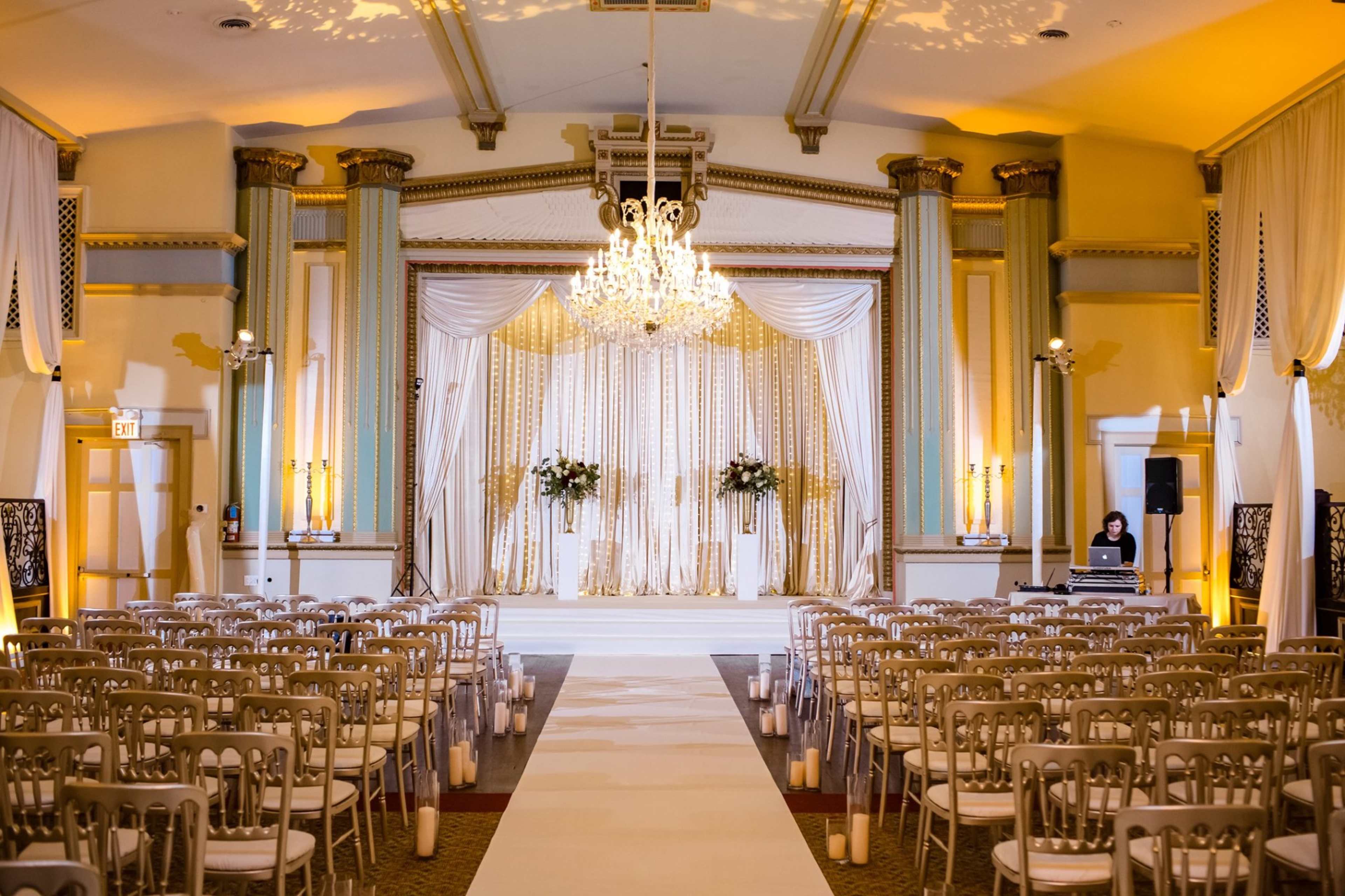 A spacious event hall is set up for a wedding ceremony, featuring rows of chairs facing a decorated altar with a chandelier above.