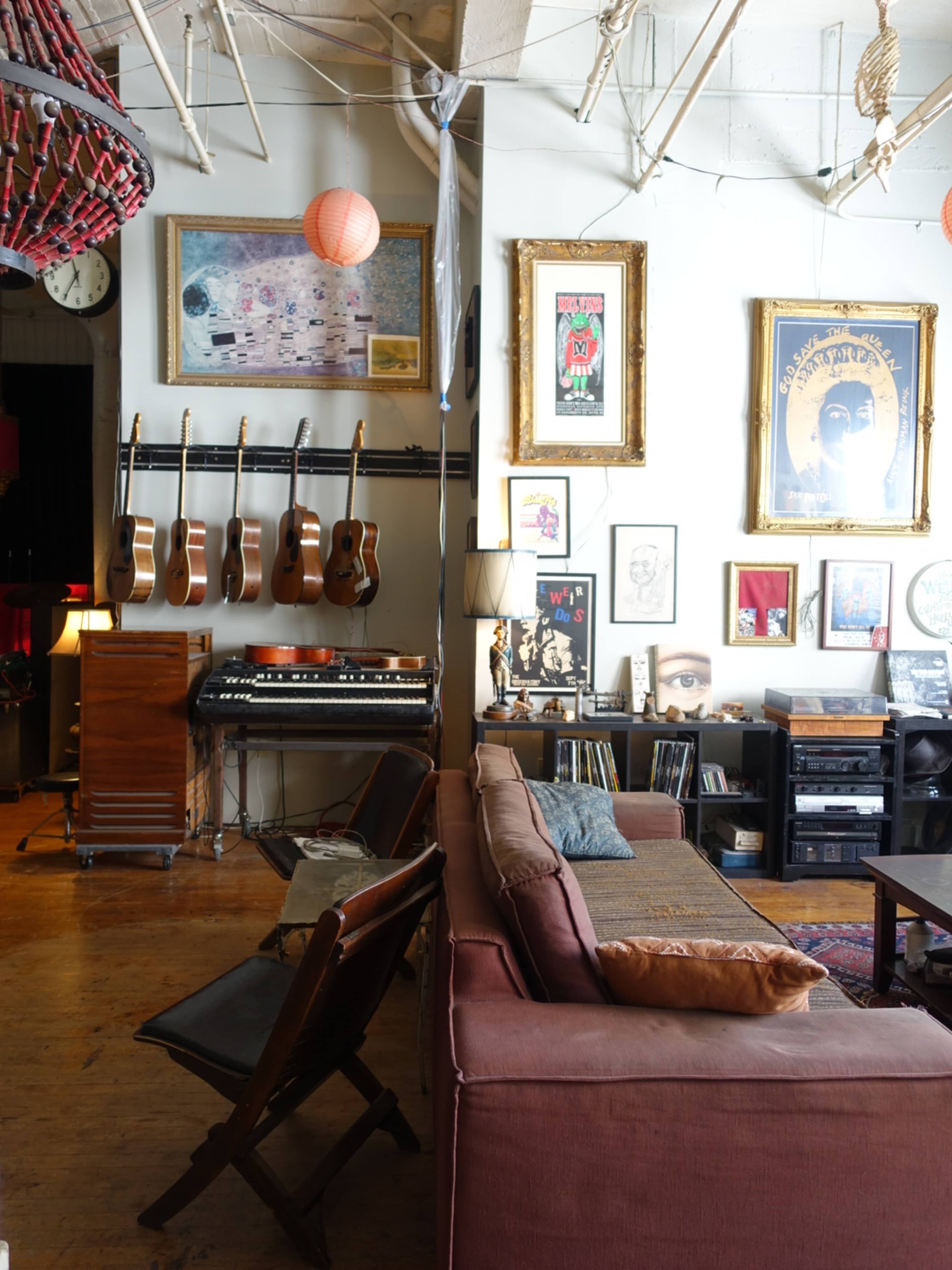 The image shows a cozy living space with a sofa, a wooden chair, several guitars on a rack, a vintage keyboard, and an array of framed artwork on the walls.